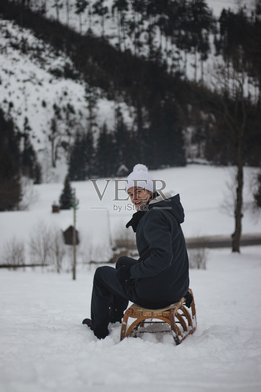 一名少年面带微笑地坐在一架历史悠久的木制雪橇上，在经过一段时间后回头看了看他设法下山的那座山。一段可怕的旅程后的快乐微笑。生活方式的隔离。新鲜的空气照片摄影图片