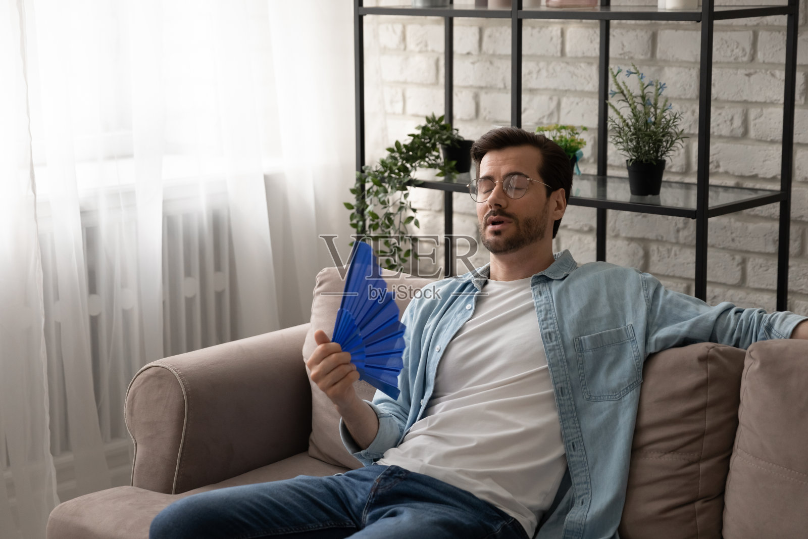 沮丧的家伙躺在沙发上用电扇降温照片摄影图片