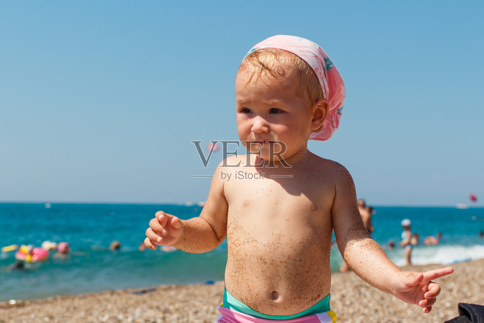 美丽的一岁小女孩穿着粉红色的泳裤站在夏天的海滩上。沙子粘在孩子身上照片摄影图片