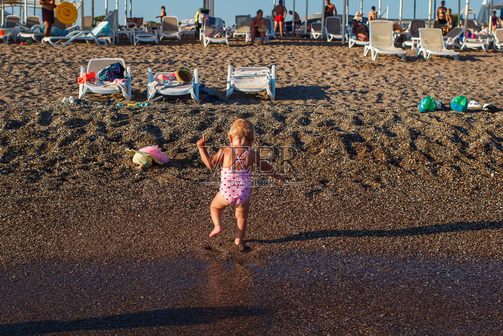 晒黑的一岁小女孩穿着粉红色泳衣，背对着镜头在沙滩上走照片摄影图片