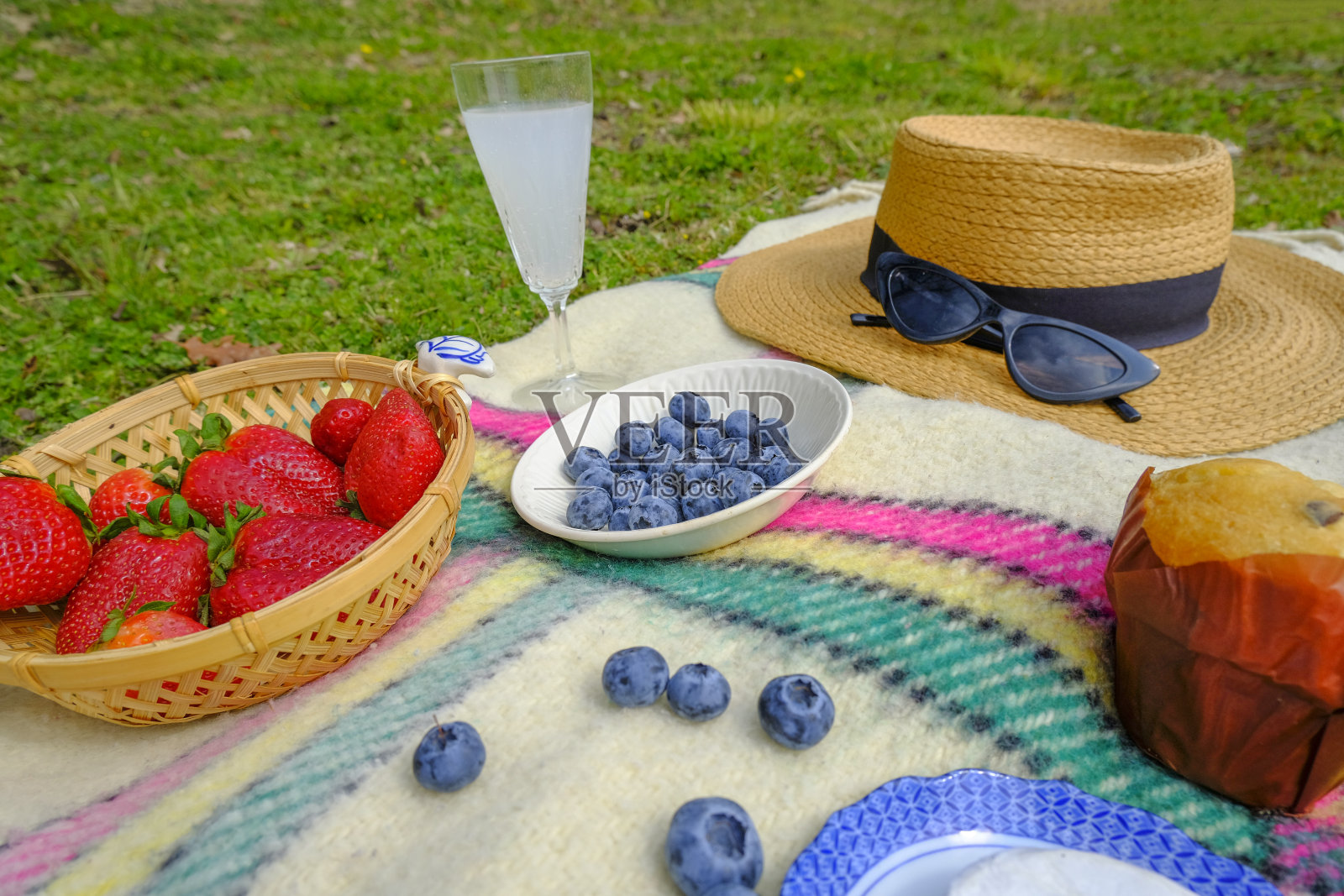 夏天在公园野餐。蓝莓盘近距离上的格子，盘子与草莓，葡萄酒杯与饮料，草帽，太阳镜横跨绿色草地。照片摄影图片