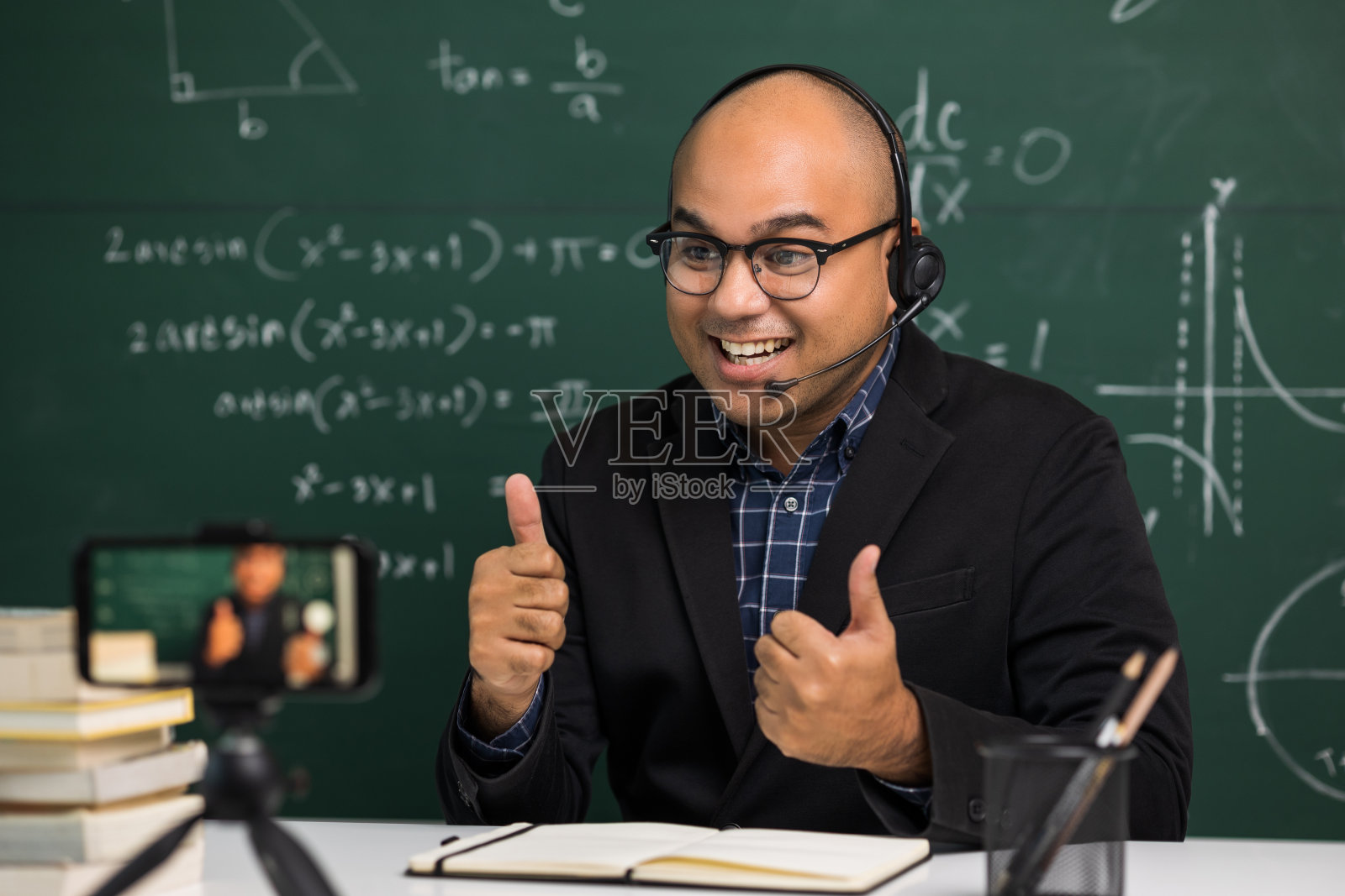 印度年轻教师男子戴着耳机坐着通过智能手机进行在线视频会议直播。亚洲数学教师在线授课，供学生在线学习。照片摄影图片