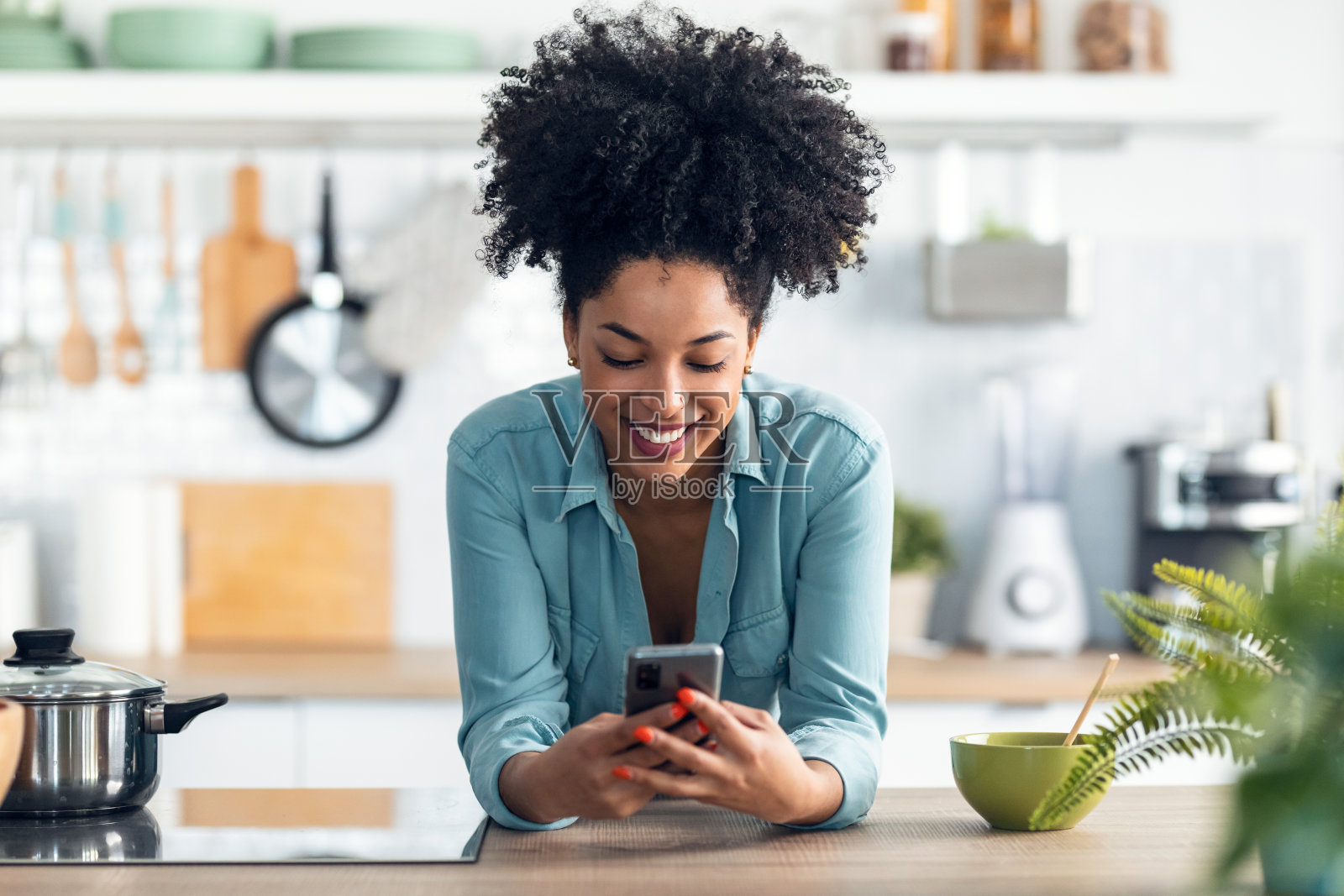美丽的非洲裔女子一边用手机发短信，一边用筷子在厨房里吃面条。照片摄影图片