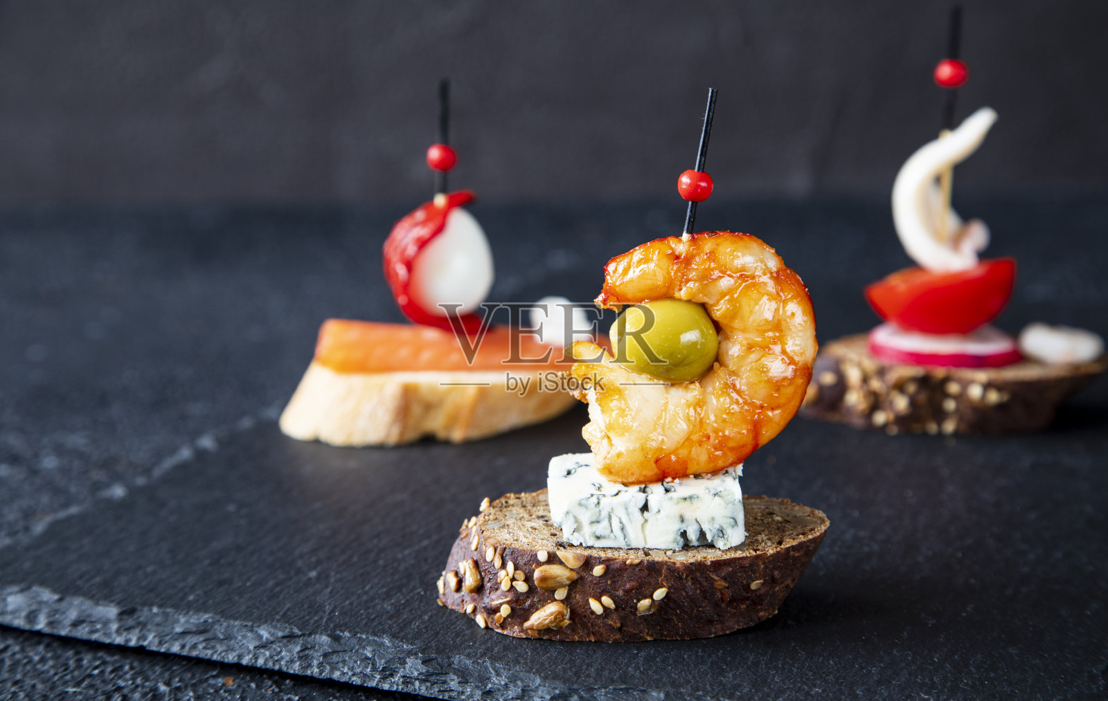 一组三餐前小吃-传统的西班牙三明治在黑板上为餐前小吃酒吧服务。三明治由高根佐拉干酪、虾、橄榄、鲑鱼、鹌鹑蛋、胡椒、萝卜、西红柿和鱿鱼组成照片摄影图片