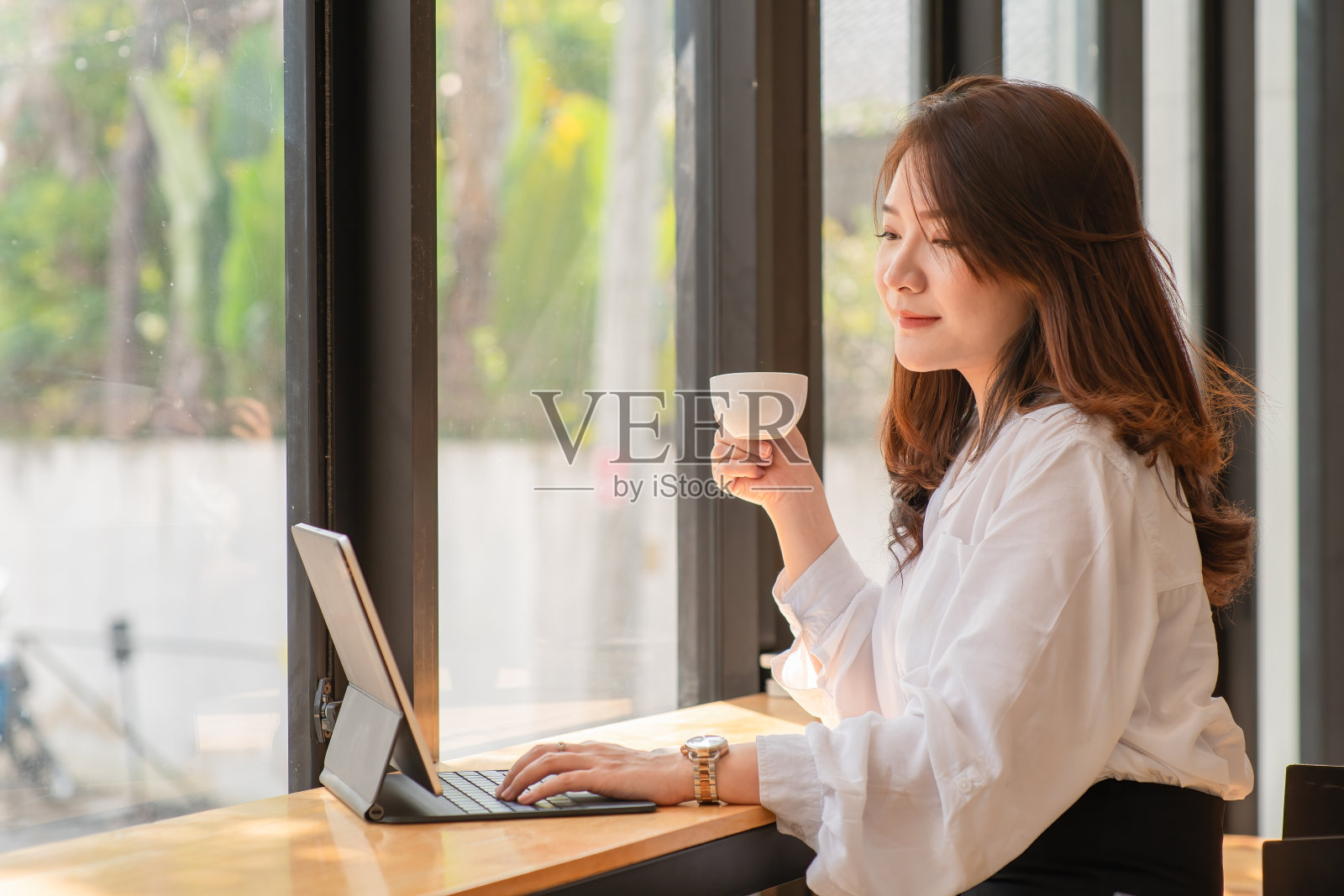 这是一幅年轻的亚洲女商人的肖像，她戴着眼镜，拿着马克杯，坐在办公桌前用平板电脑工作。照片摄影图片