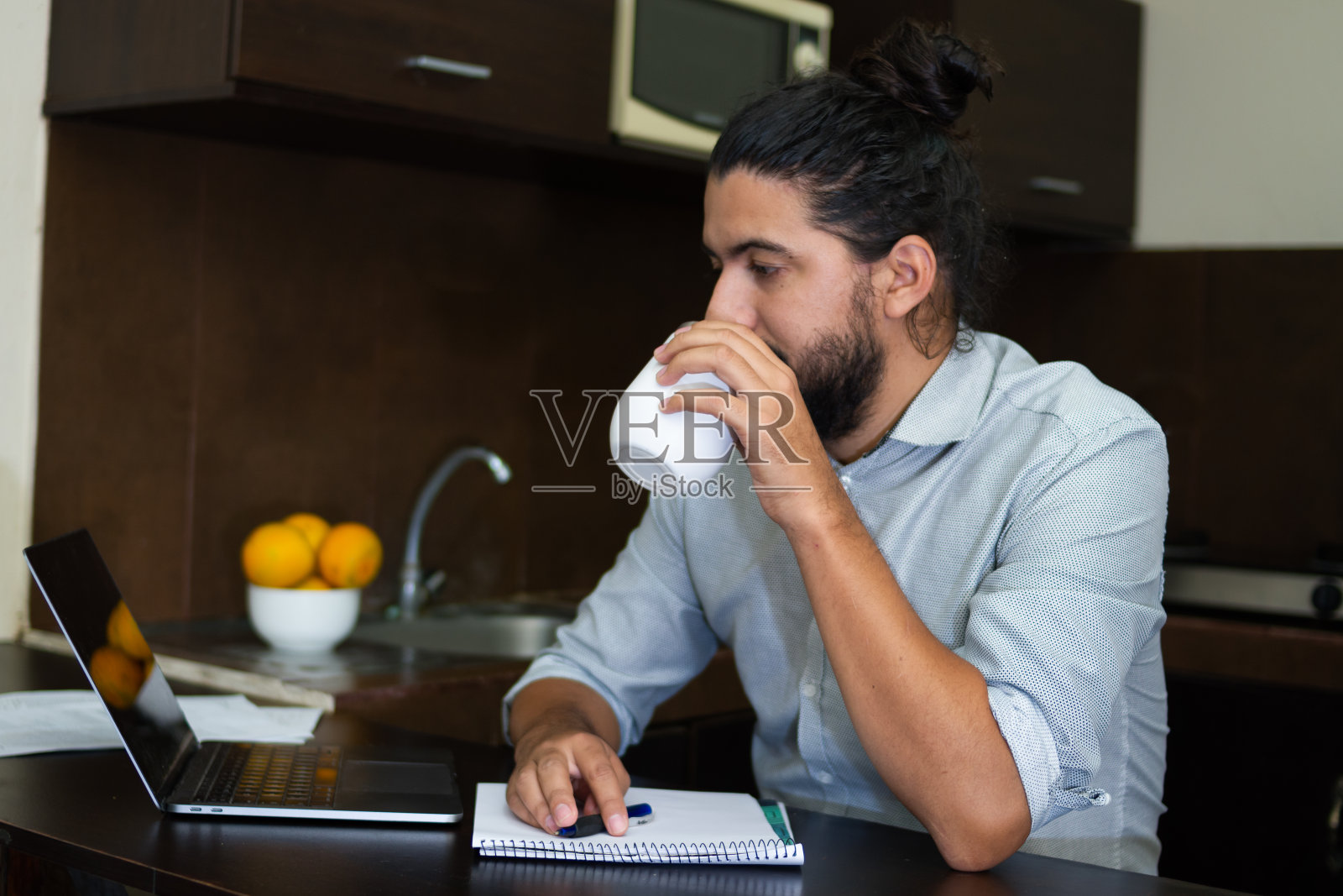 年轻人放松地坐在厨房里喝着饮料，看着笔记本旁边的笔记本电脑，在家里工作。照片摄影图片