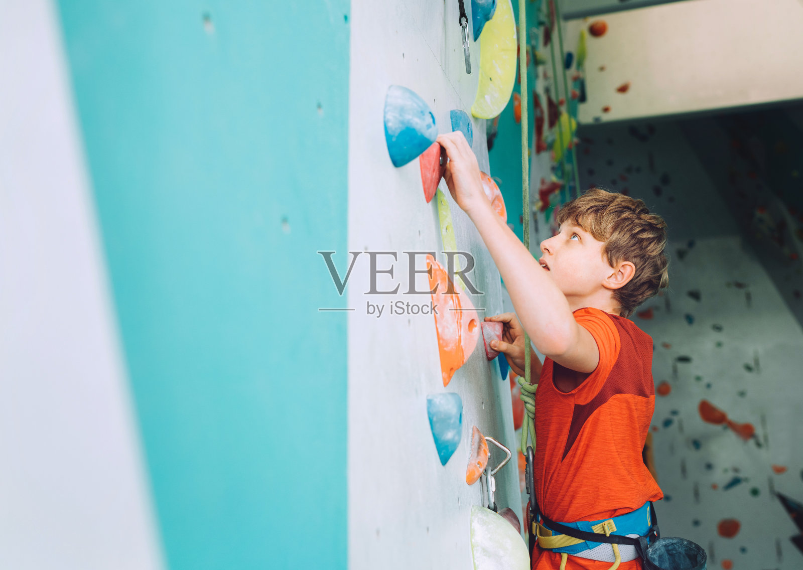 微笑的少年在室内攀岩墙大厅。这个男孩正在用顶绳和攀爬挽具攀爬。活跃的青少年时间消费概念形象照片摄影图片