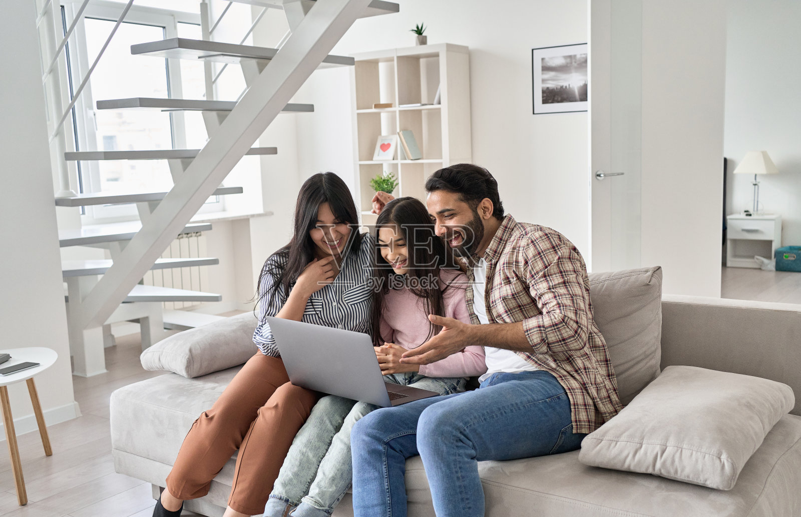 快乐的印度家庭与女儿在家中使用笔记本电脑玩耍。微笑的父母和十几岁的孩子笑着进行虚拟视频通话会议，坐在沙发上看有趣的在线电视照片摄影图片