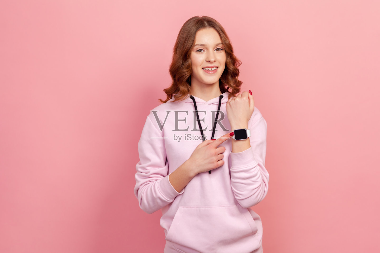 一幅欢快的年轻褐发女人的肖像，穿着帽衫指着手表，微笑着，显示约定的见面时间，快乐的分钟照片摄影图片