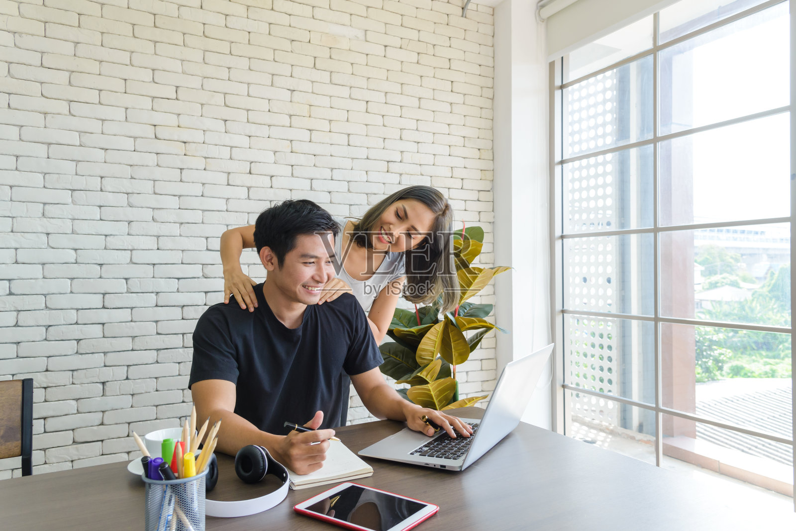 年轻的亚洲夫妇穿着休闲的衣服，坐在现代厨房的大木桌和工作与电脑笔记本电脑在家里。爱情、幸福、在家工作的概念照片摄影图片