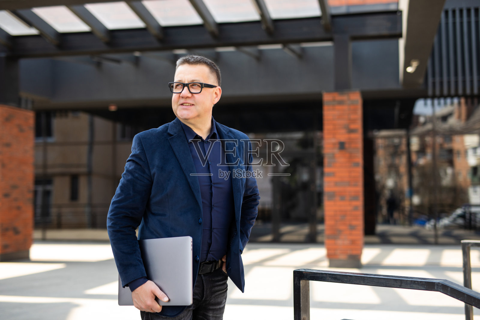 英俊成熟的商人戴着时髦的眼镜，拿着笔记本电脑，站在大街上。成功的经营理念照片摄影图片