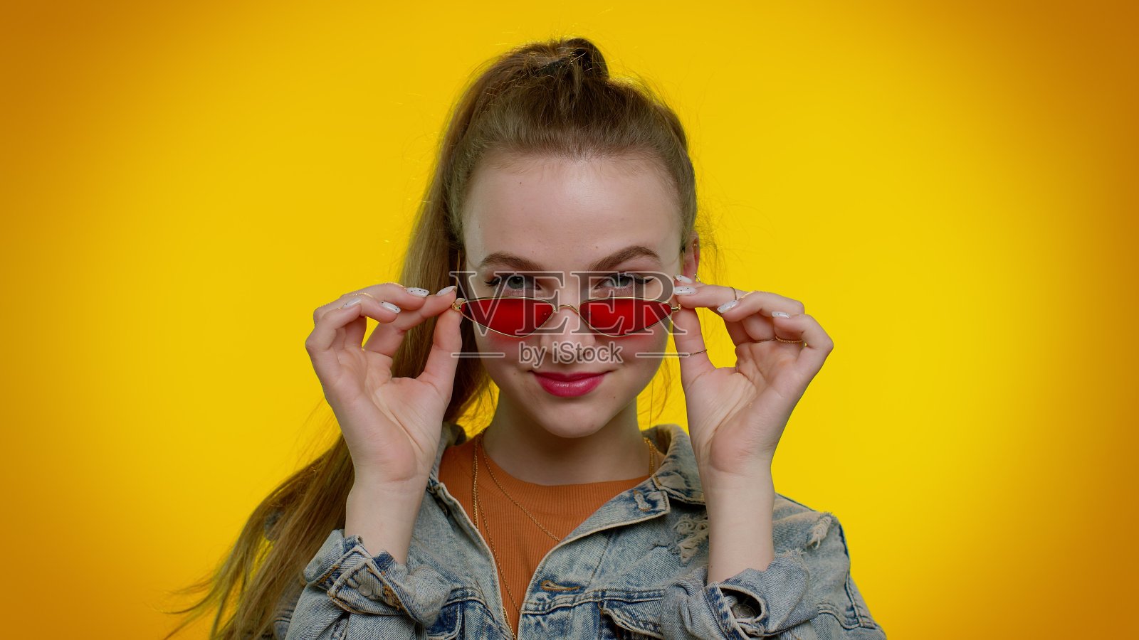 性感开朗的时尚女孩穿着牛仔夹克戴着太阳镜，迷人的微笑在黄色的墙上照片摄影图片