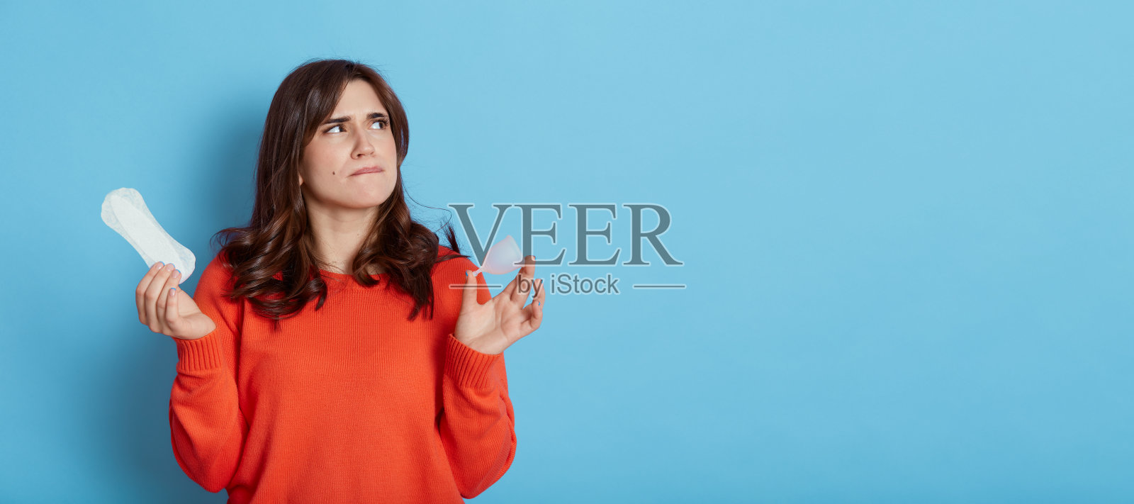 沉思美丽的女性穿着休闲橘色毛衣看向一边，带着体贴的面部表情，手持月经杯和垫，广告拷贝空间，孤立在蓝色背景上。照片摄影图片