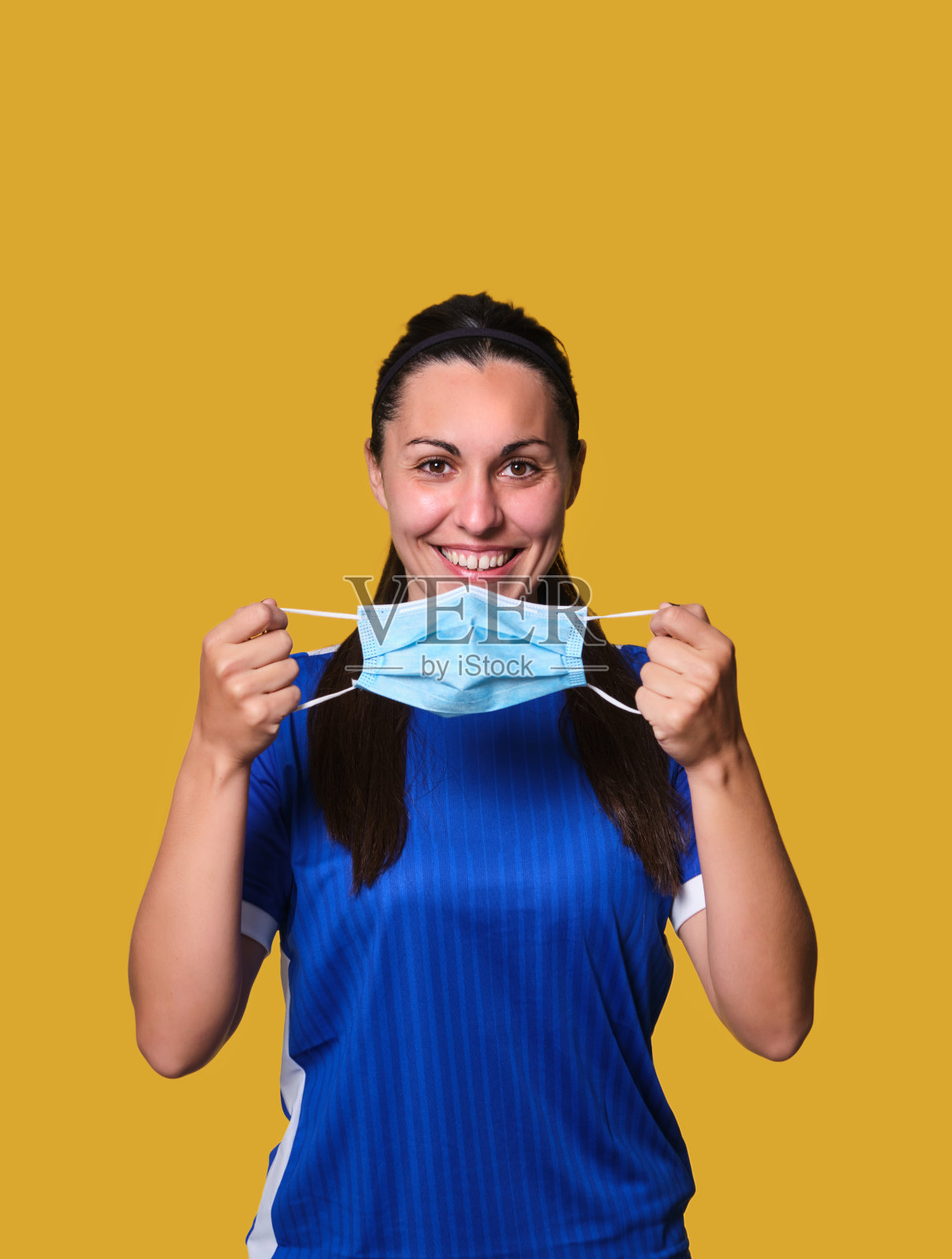 年轻女足球运动员戴着防冠状病毒防护口罩，背景为黄色隔离照片摄影图片