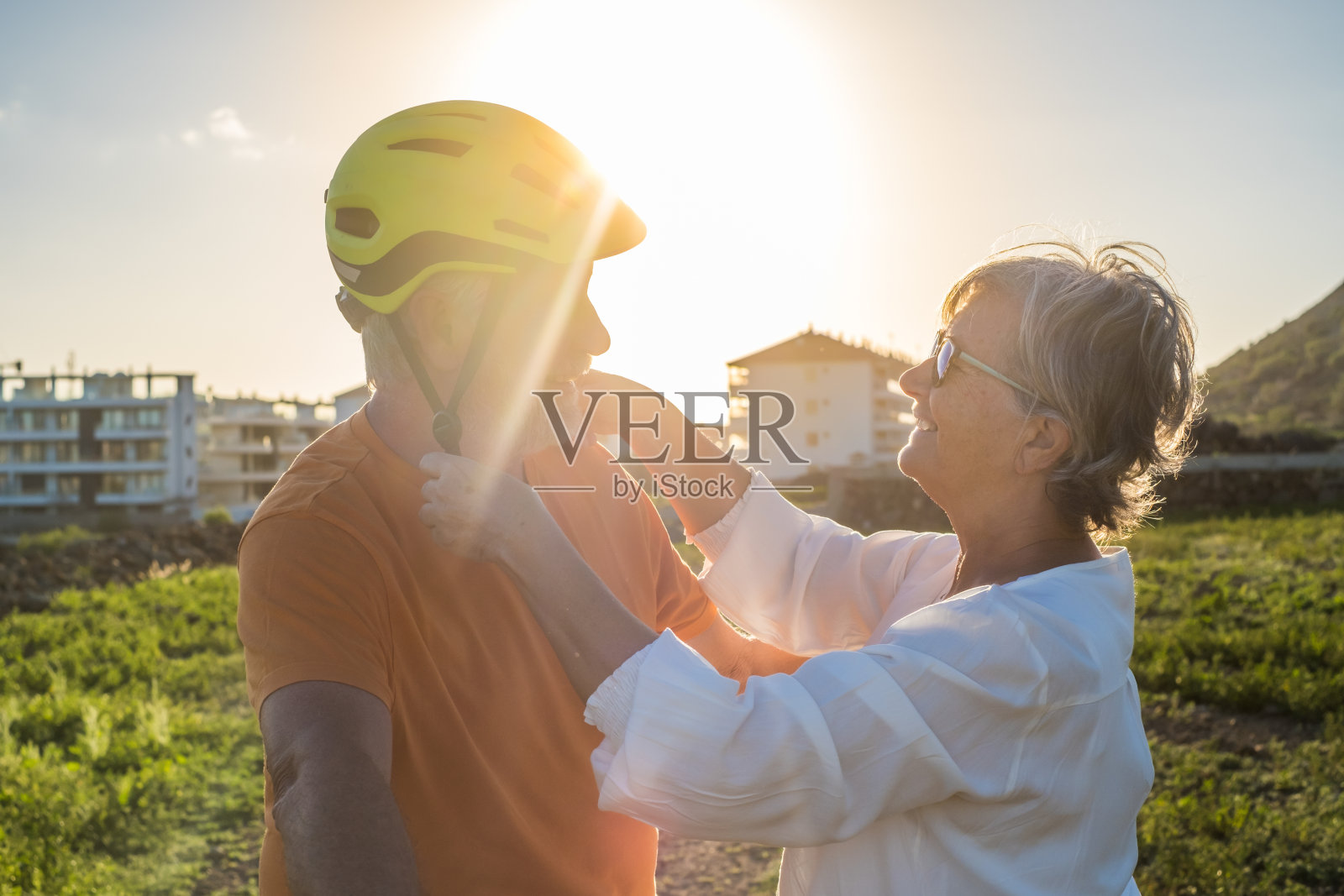 老女人给丈夫戴上防护帽骑自行车。明亮的日光照片摄影图片