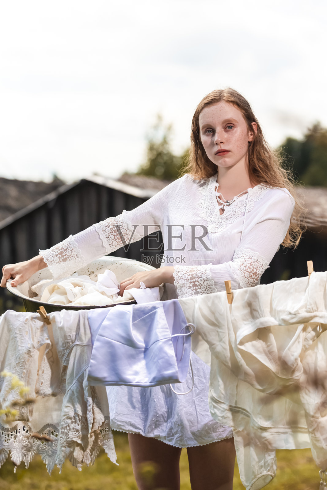 冷静的红发白人女孩与洗衣盆摆姿势的自然肖像，同时将其挂在户外的绳子在农村环境。垂直拍摄照片摄影图片