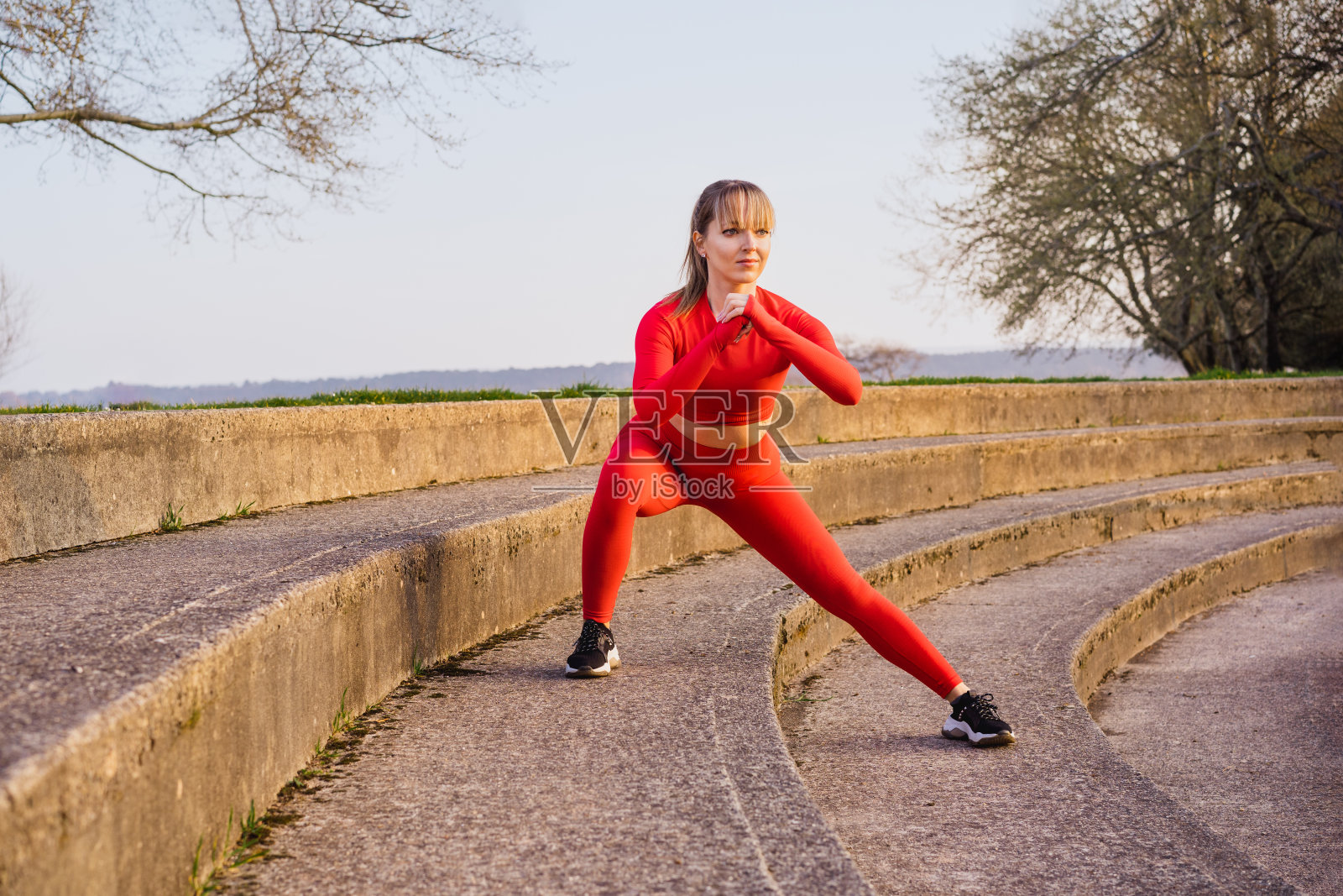 年轻的健身妇女在红色运动服做侧弓步，蹲在户外运动在体育场的台阶上。跑步后做伸展运动。积极的生活方式和保健。副本的空间。照片摄影图片