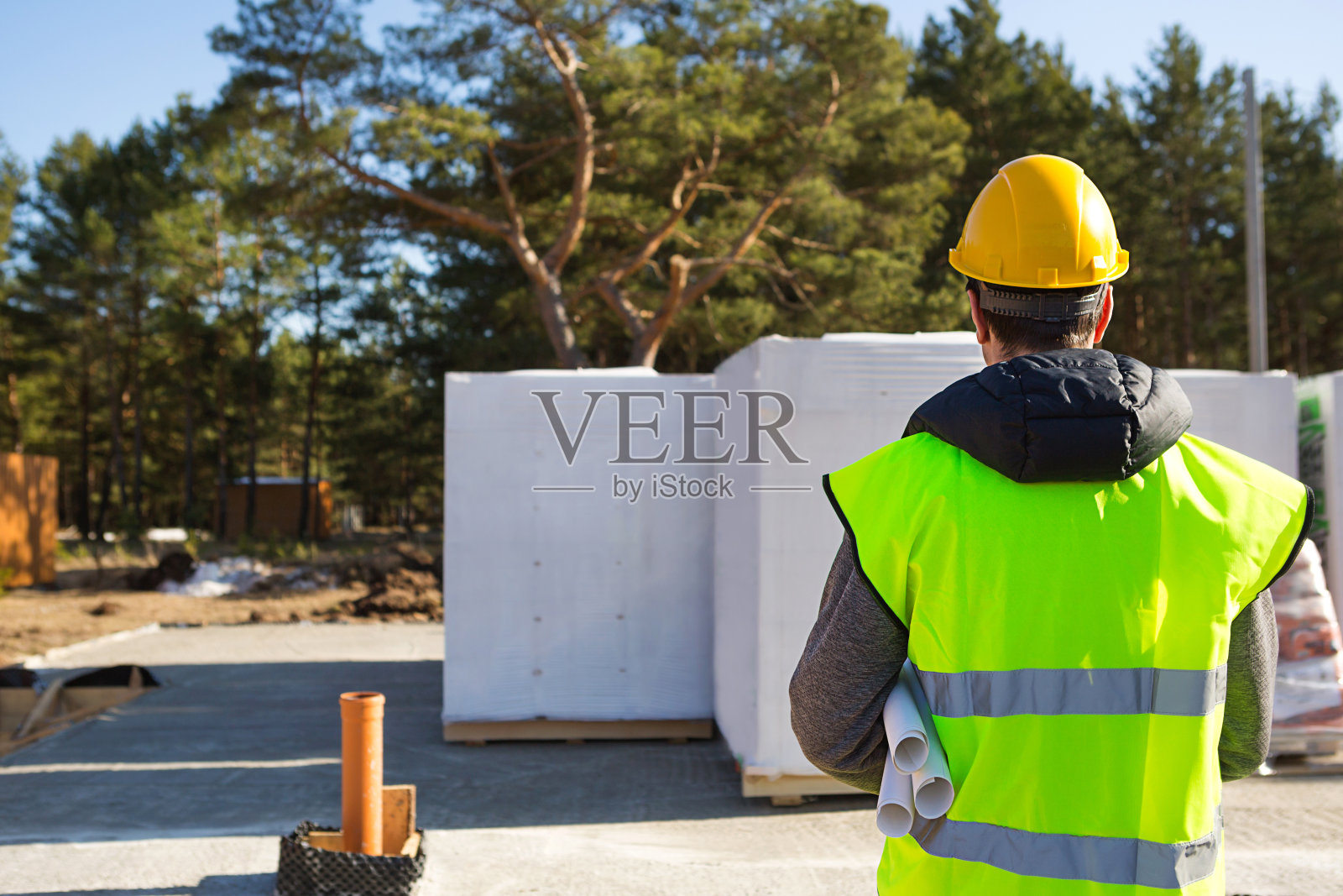 该项目建筑师的后背是一所房子的施工现场，奠定了基础，交付了砌块。建筑工人戴着黄色安全帽和信号背心。模拟了照片摄影图片