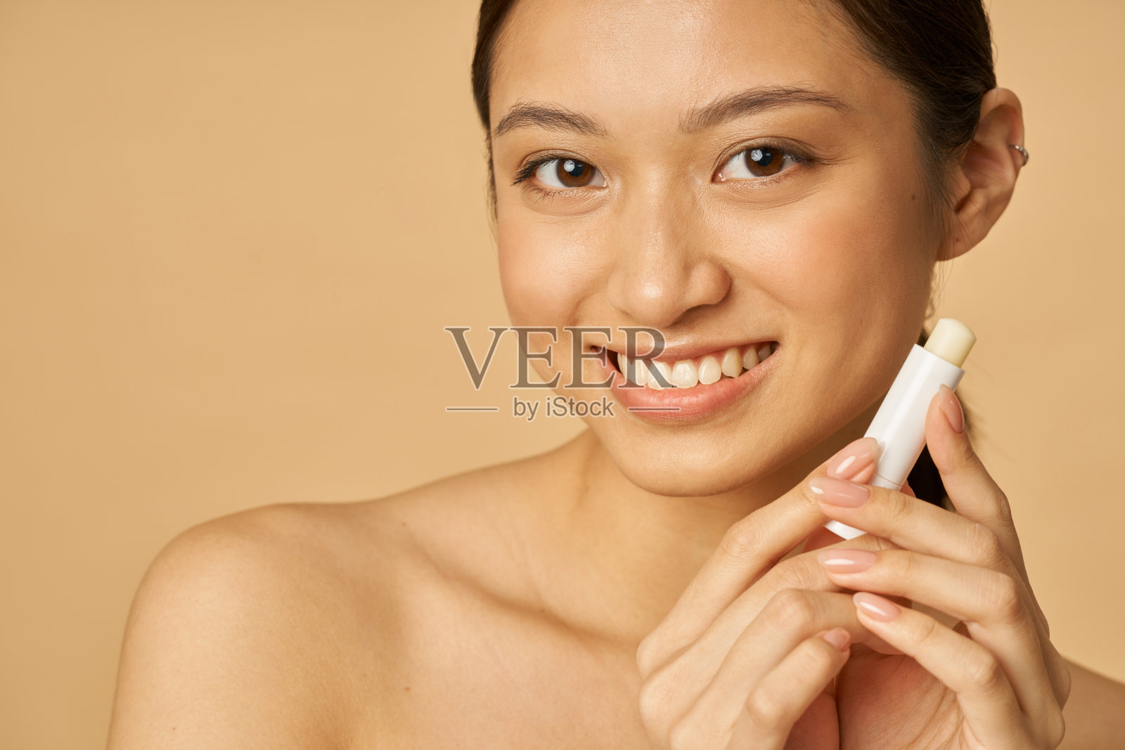 可爱的年轻女子微笑的近距离肖像，而持有唇膏，摆出孤立的米色背景照片摄影图片