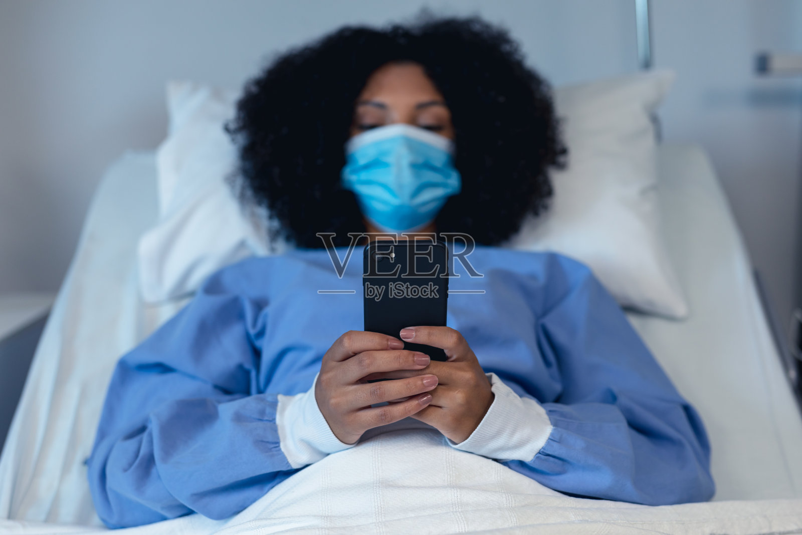 非裔美国女性患者戴着口罩躺在医院病床上使用智能手机照片摄影图片