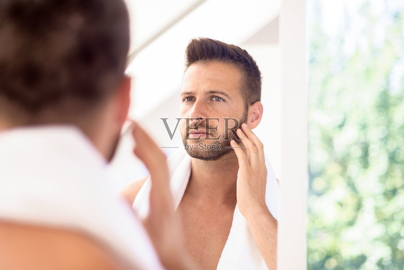 英俊的男人站在浴室镜子前，看着自己的脸照片摄影图片