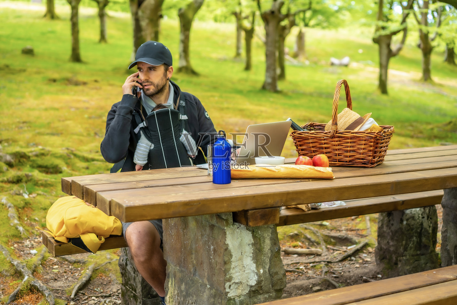 一个年轻的白种父亲坐在野餐桌边远程工作，电脑，孩子在背包里。夏天的生活方式照片摄影图片