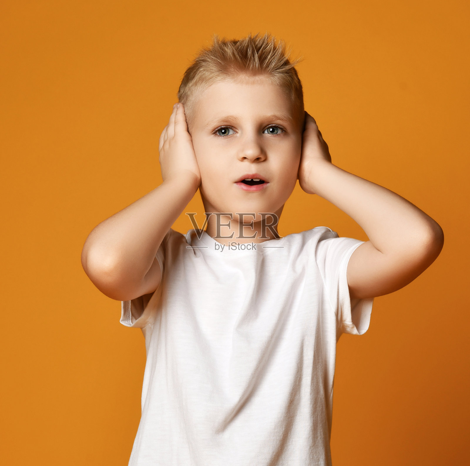 金发男孩穿着白色t恤和牛仔裤站着，用手捂住耳朵照片摄影图片