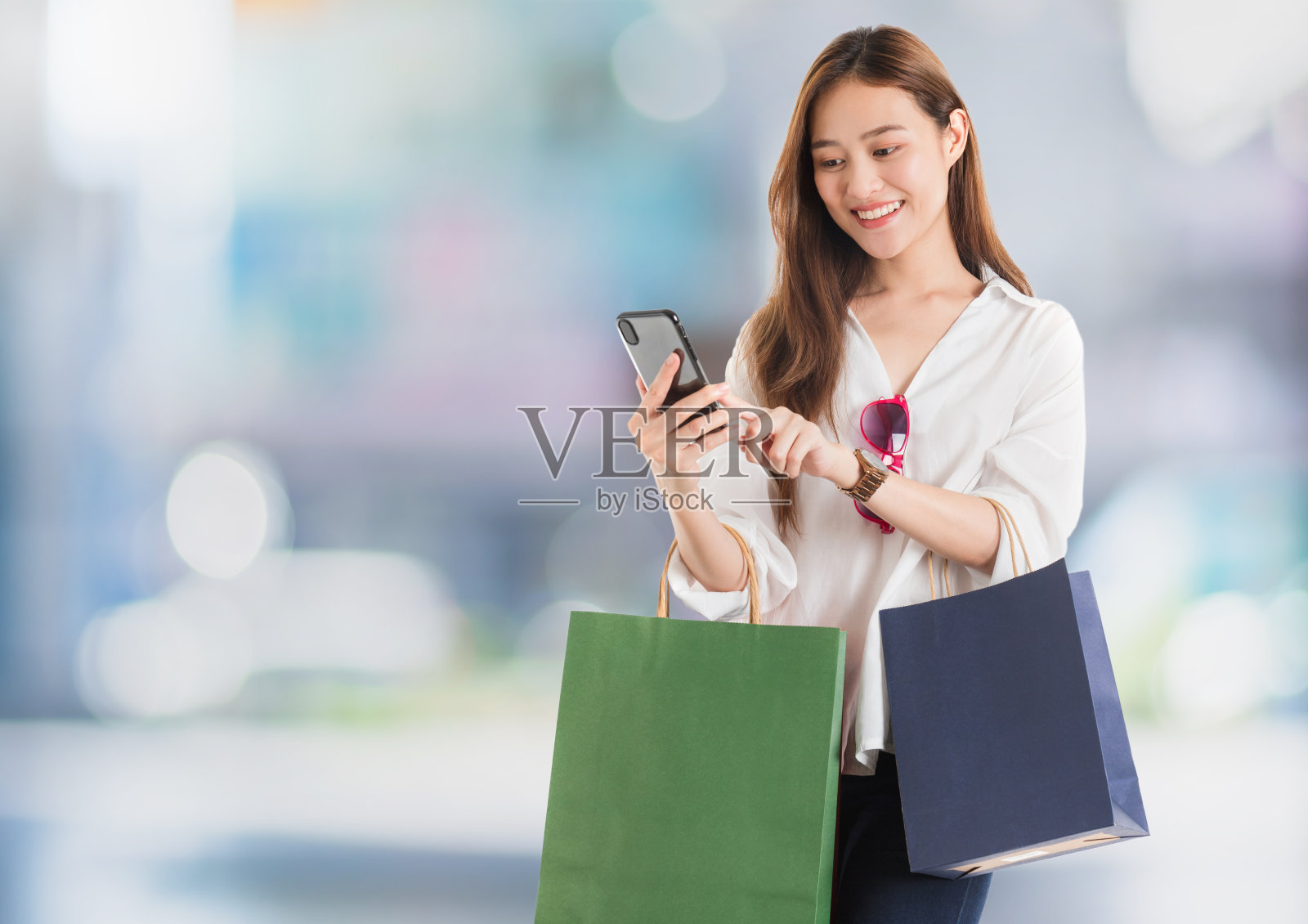 亚洲美女博主正在用智能手机在线购物，购物袋在模糊的商场内部背景中。网上购物业务的概念。照片摄影图片