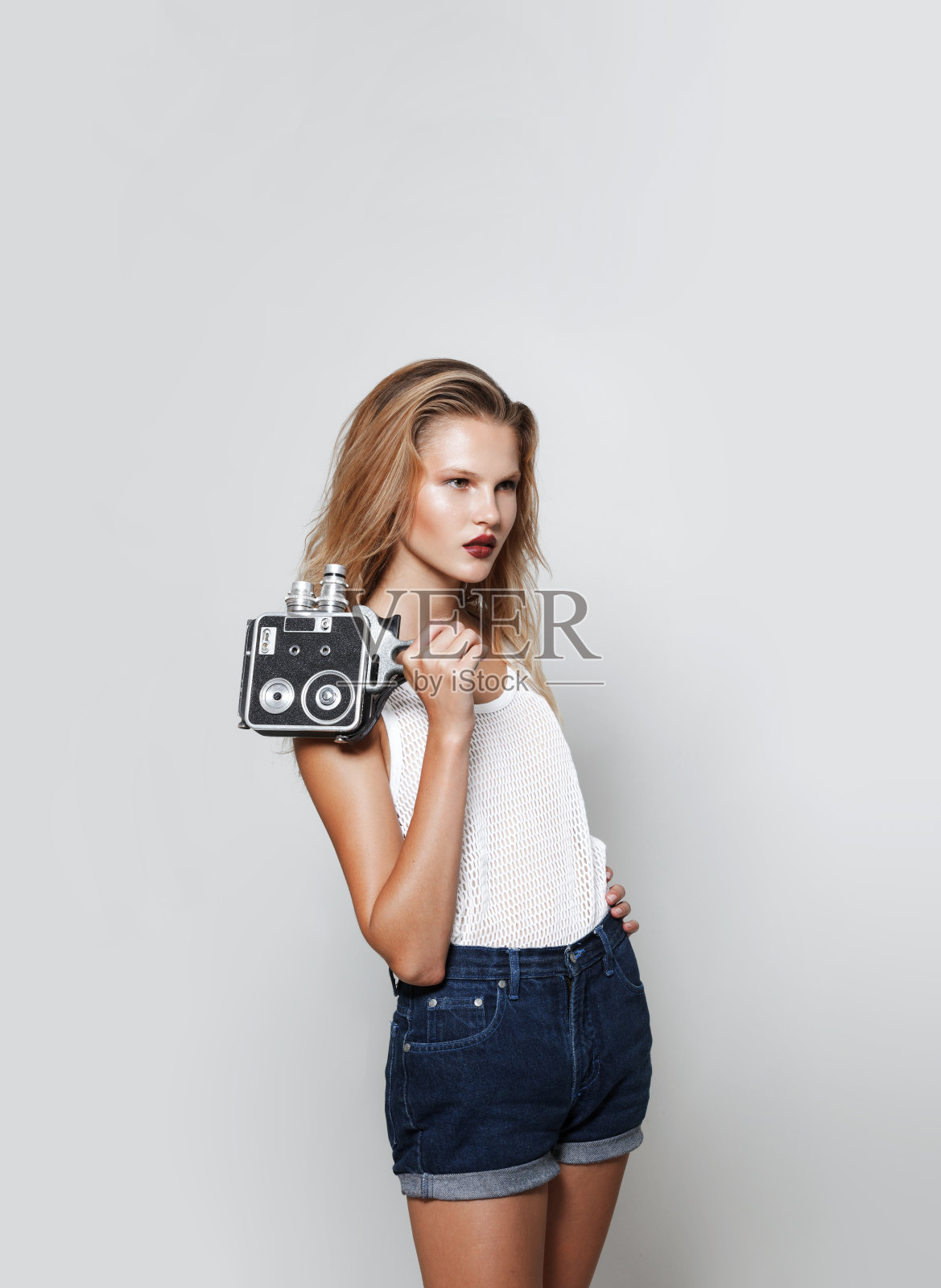 年轻女人的肖像金发碧眼摆姿势在工作室白色时尚衣服，看着别处，拿着一个复古相机在手里。照片摄影图片