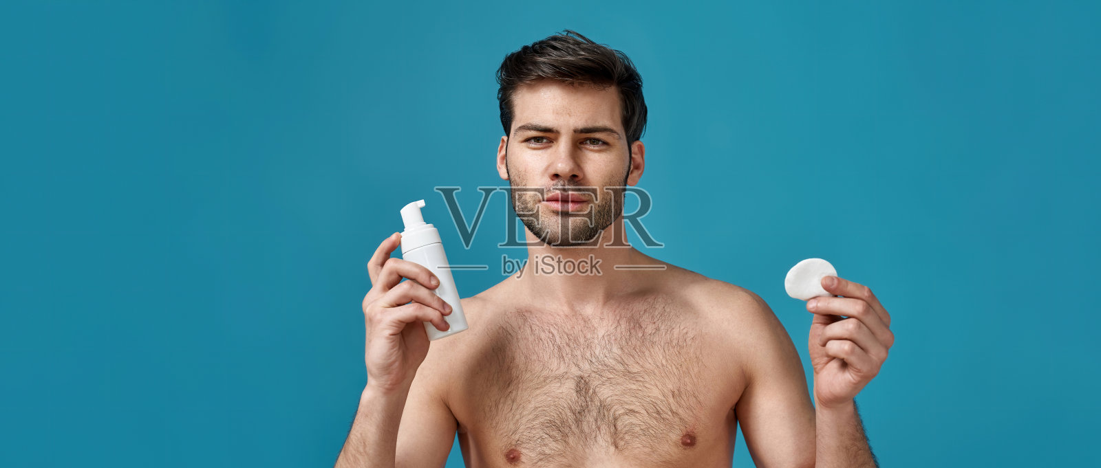 英俊的赤膊男子与深色头发拿泡沫美容产品和棉垫清洁他的脸，摆出孤立的蓝色背景照片摄影图片