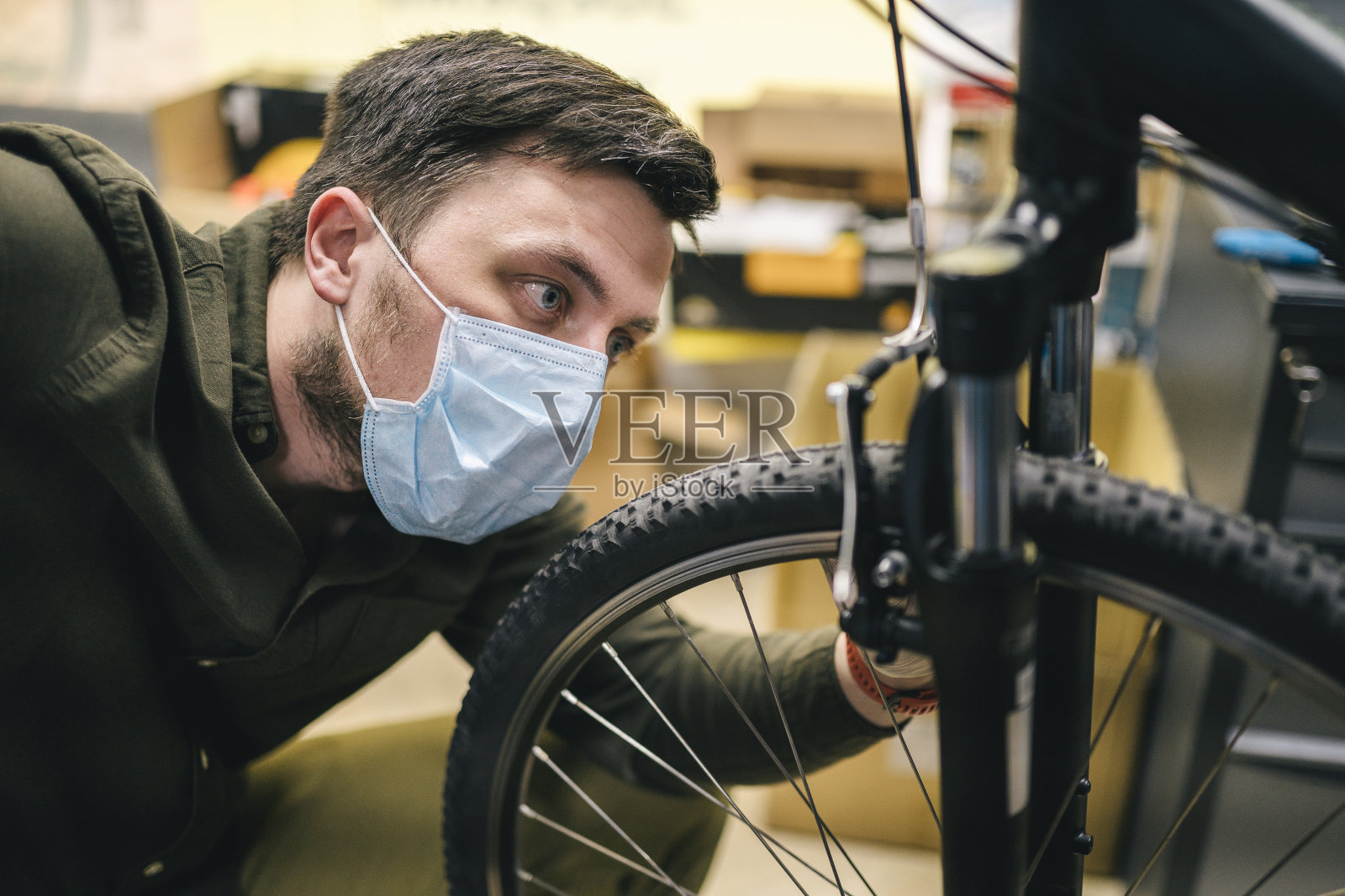 机修工戴着口罩在车间修理自行车。自行车店工作人员戴着防护口罩在隔离期间修理自行车。修理工维修周期。体育商店的概念照片摄影图片