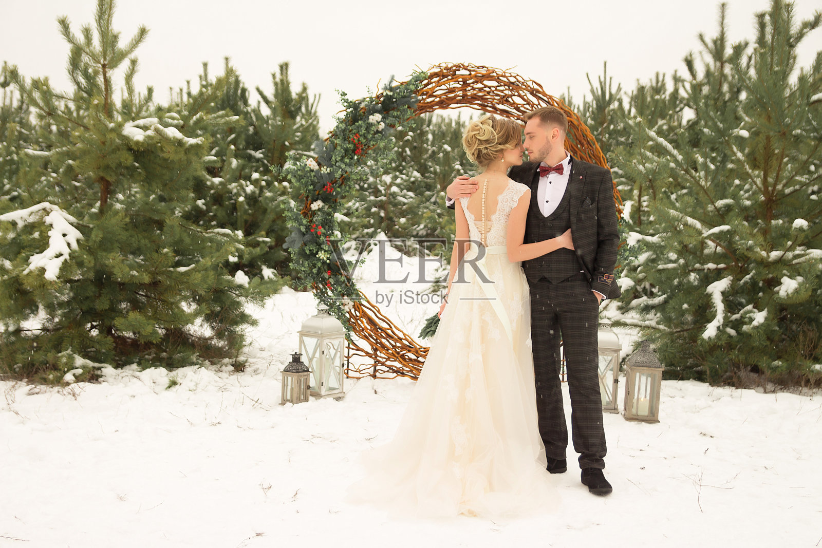 一对夫妇，一男一女，正站在一片冬季森林的背景下，周围有一圈装饰着藤蔓的鲜花，一件白色的连衣裙和套装。新娘和新郎。束云杉。女孩后视图照片摄影图片