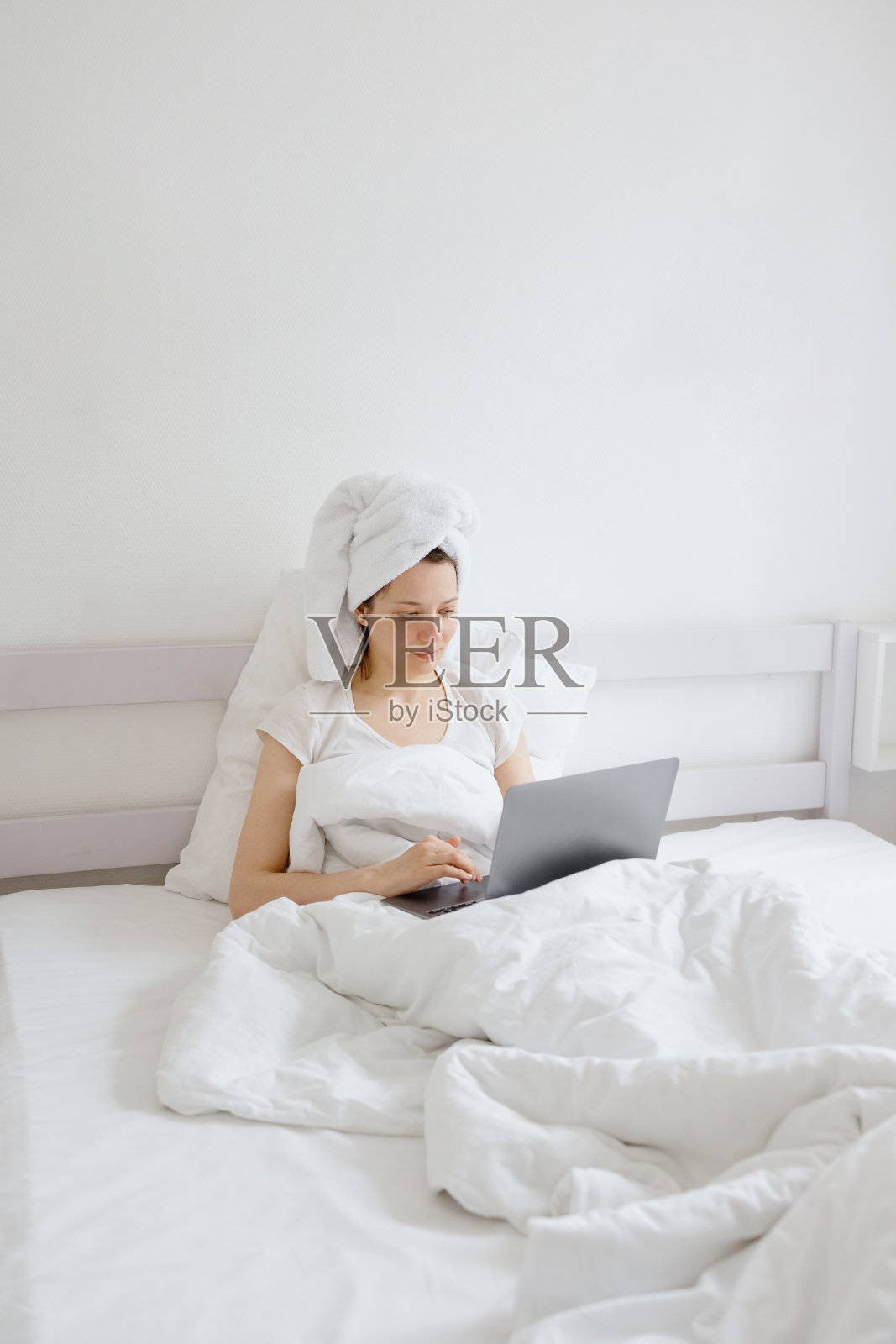 迷人的欧洲女人坐在白色的床上，头发上放一条毛巾，在她的手机或笔记本电脑上聊天，在度假或旅游中放松照片摄影图片