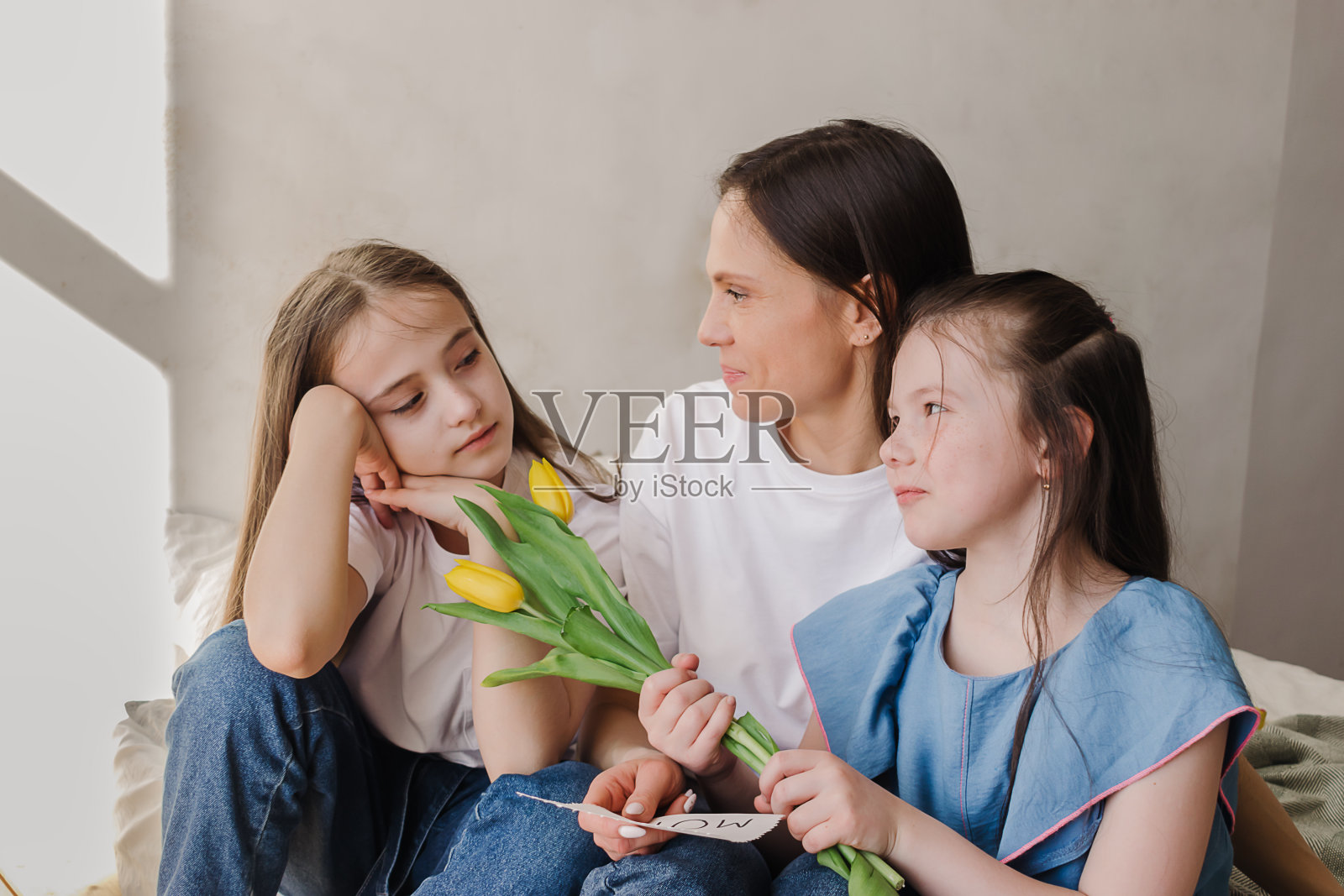 微笑的黑发年轻女子与她的女儿在家里交谈。她手里拿着一束黄色的花和自制的贺卡。母亲节、3月8日和生日的概念照片摄影图片