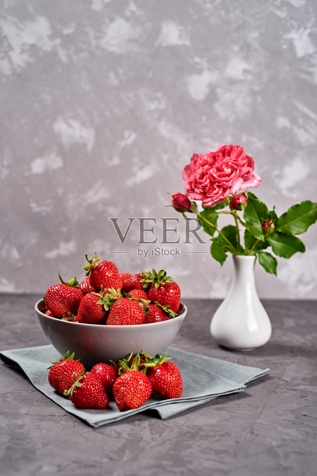 成熟的草莓在陶瓷碗上亚麻餐巾和粉红色的玫瑰花瓶在灰色的混凝土背景，复制空间。健康食品理念，静物生活照片摄影图片