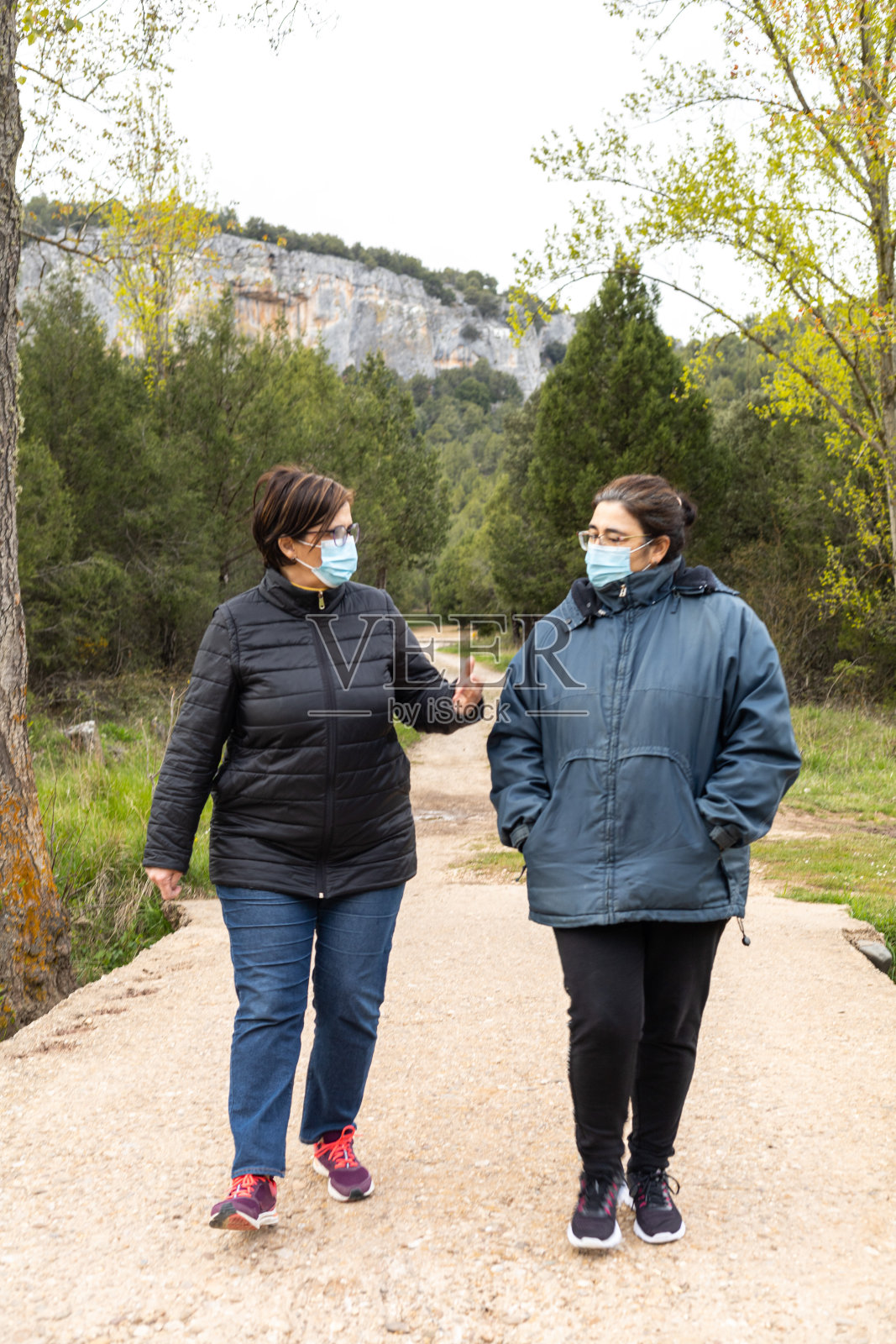 在阳光明媚的日落和山景中，两名中年妇女戴着2019冠状病毒口罩行走在乡间小路上。照片摄影图片