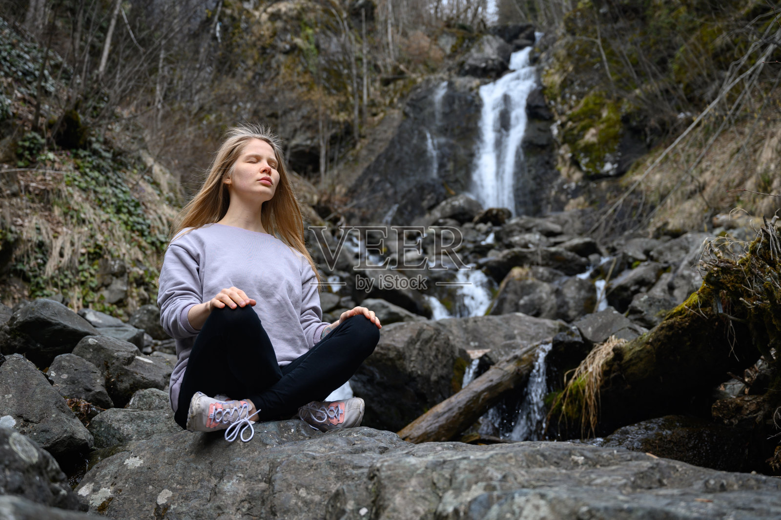 女孩坐在一块大石头上，在山河旁边的瀑布旁冥想。照片摄影图片