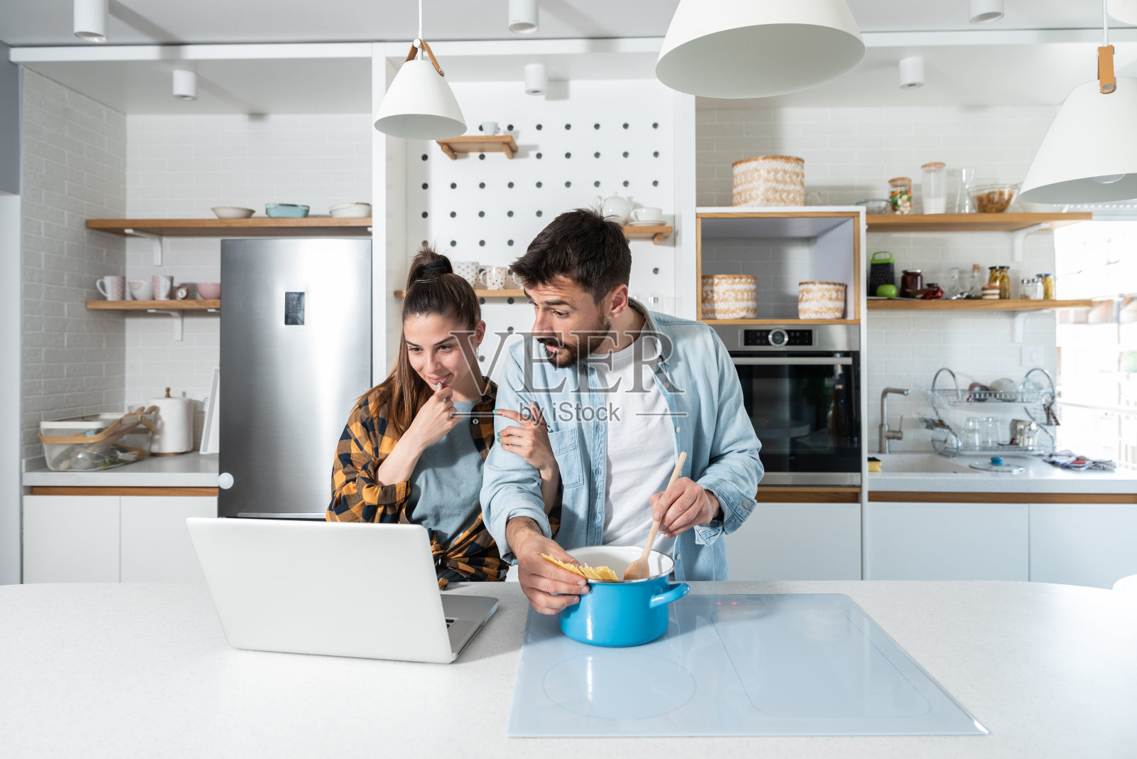 年轻夫妇在他们的厨房里烹饪，通过在他们的笔记本电脑上观看在线课程准备食物照片摄影图片