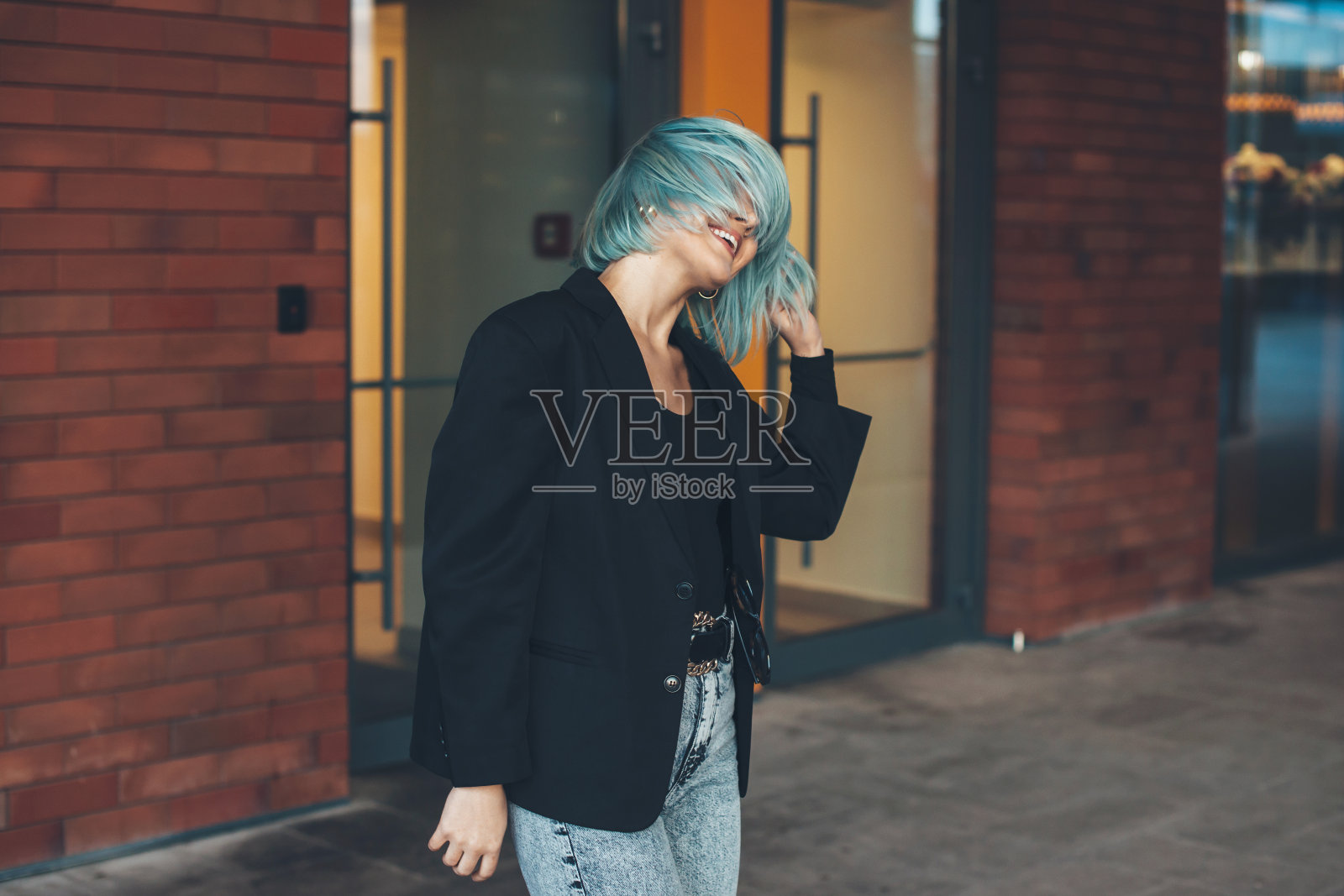 一个时髦的蓝头发的女人在城市的一堵砖墙附近摸着她的头发笑着照片摄影图片