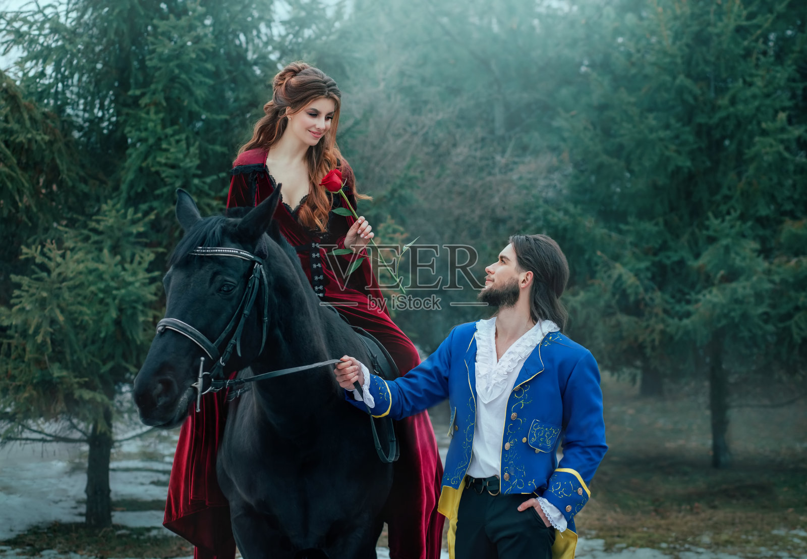 中世纪爱情情侣：男子在冬日森林中赠与女子玫瑰，复古红色长裙和蓝色外套，王子与公主，黑马，红发照片摄影图片