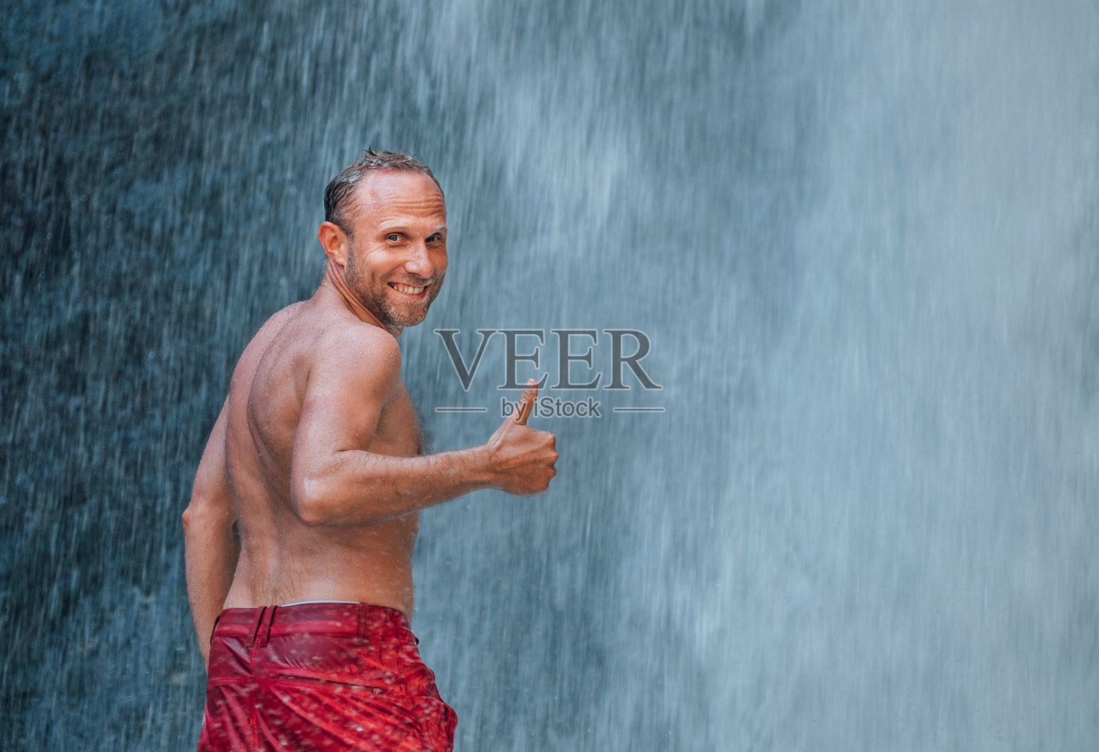 中年赤裸上身真诚地笑男人站在山河瀑布下显示大拇指!大自然的力量。适合人们，徒步旅行，并具有自然美的概念形象。照片摄影图片