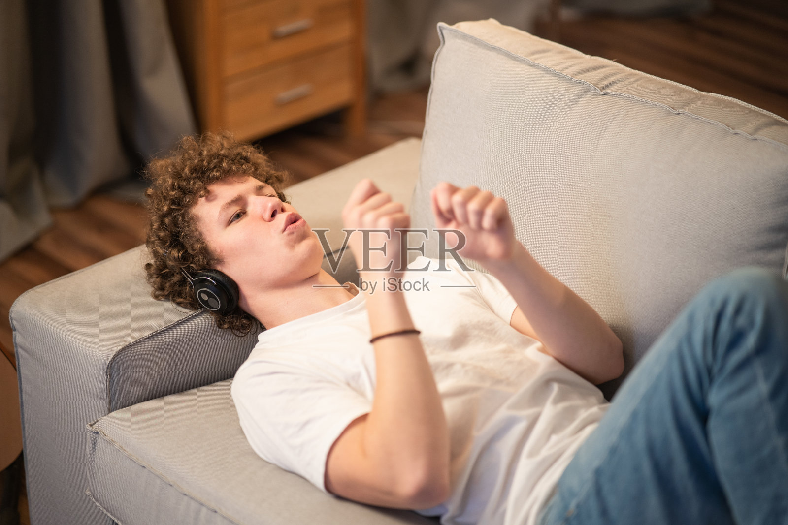 一个卷发的小男孩躺在家里的沙发上听音乐，他最喜欢的乐队，一首有趣的歌。这位少年戴着无线耳机跳舞、挥舞手臂、唱歌照片摄影图片