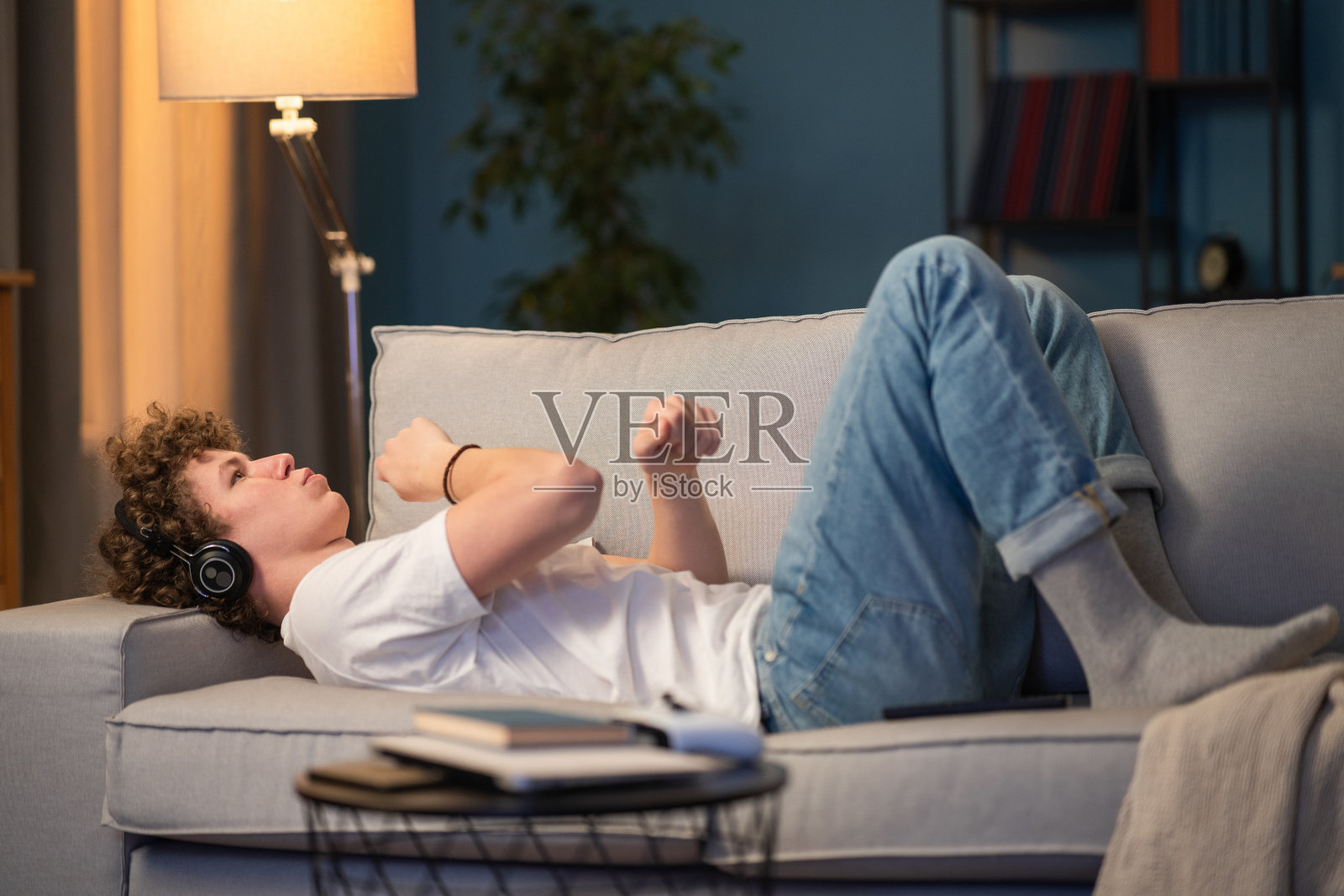 一个卷发的小男孩躺在家里的沙发上听音乐，他最喜欢的乐队，一首有趣的歌。这位少年随着音乐的节奏躺在沙发上，一边舞动着双臂照片摄影图片