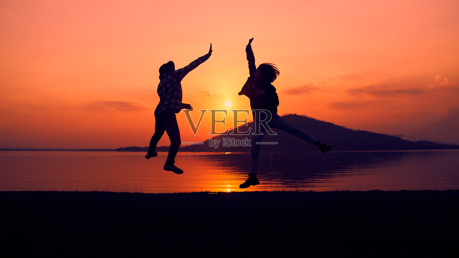夕阳西下，两个人在湖边欢快跳跃的剪影。照片摄影图片