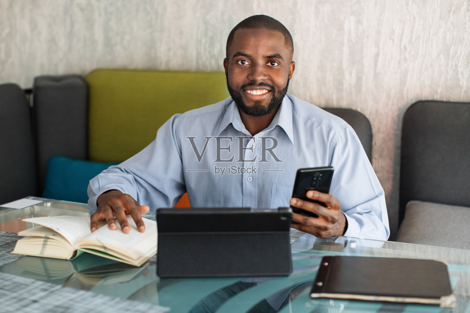 笑容满面的非洲男人，学生或自由职业者，留着胡子，穿着灰色衬衫，坐在咖啡馆或联合办公空间的桌子旁，从书本、数字平板电脑和手机中寻找信息。照片摄影图片