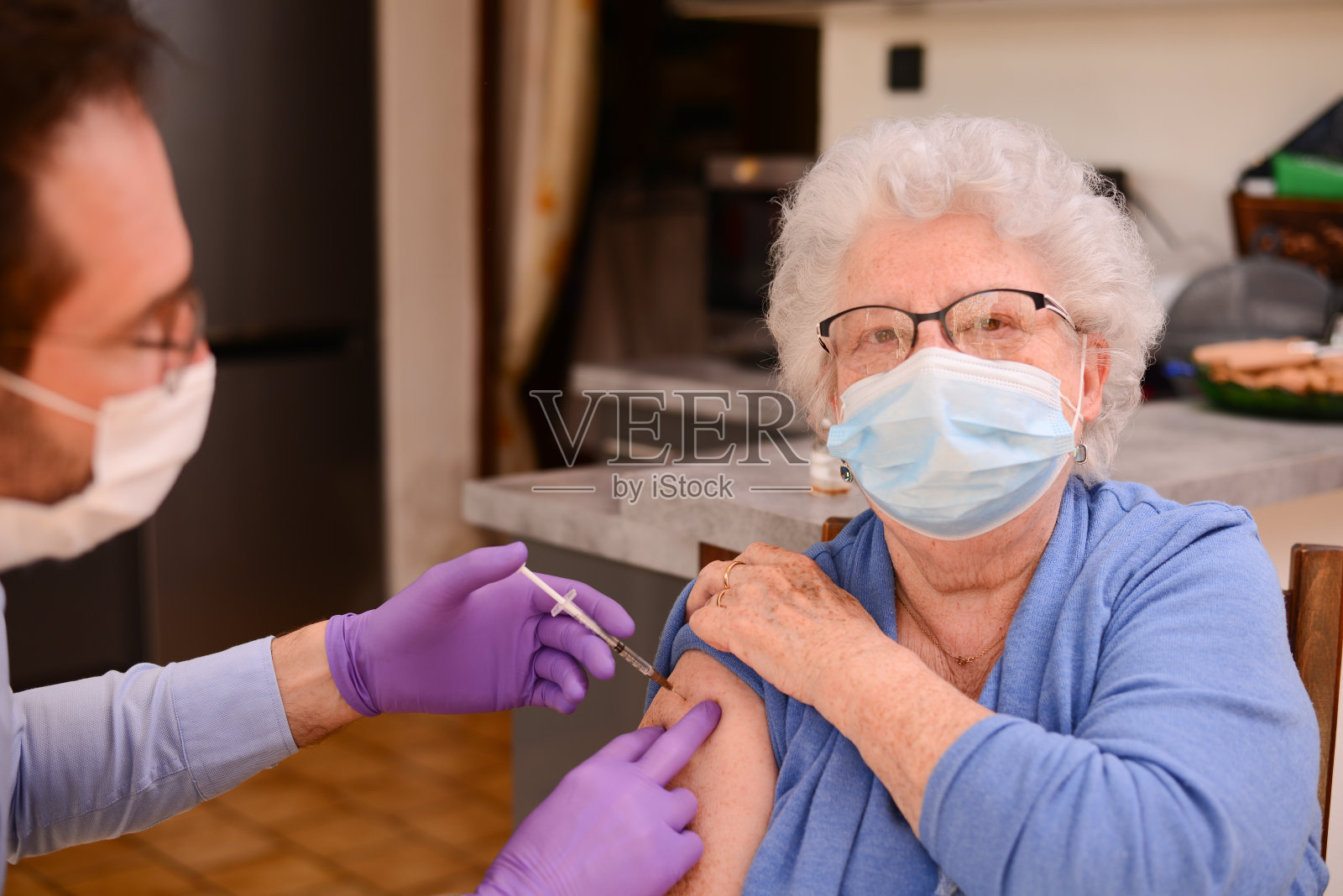 戴外科口罩的老年妇女，由男医生护士在家注射新冠肺炎疫苗照片摄影图片