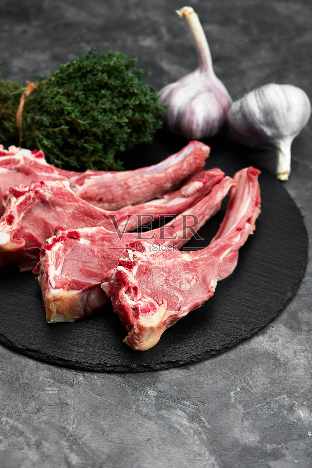 生羊排配大蒜和香草，放在黑色陶瓷盘子上，白石灰色的桌子上。前视图。与复制空间照片摄影图片