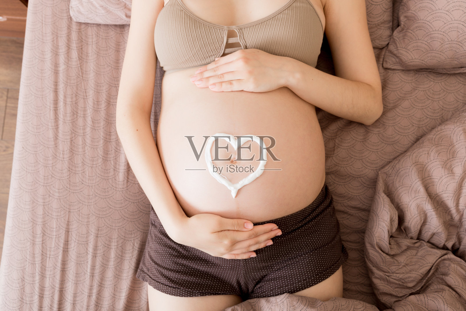 一个怀孕的女孩坐在家里的床上，在她的肚子上涂抹一种抗妊娠纹霜。怀孕、母性、准备和期望的概念照片摄影图片
