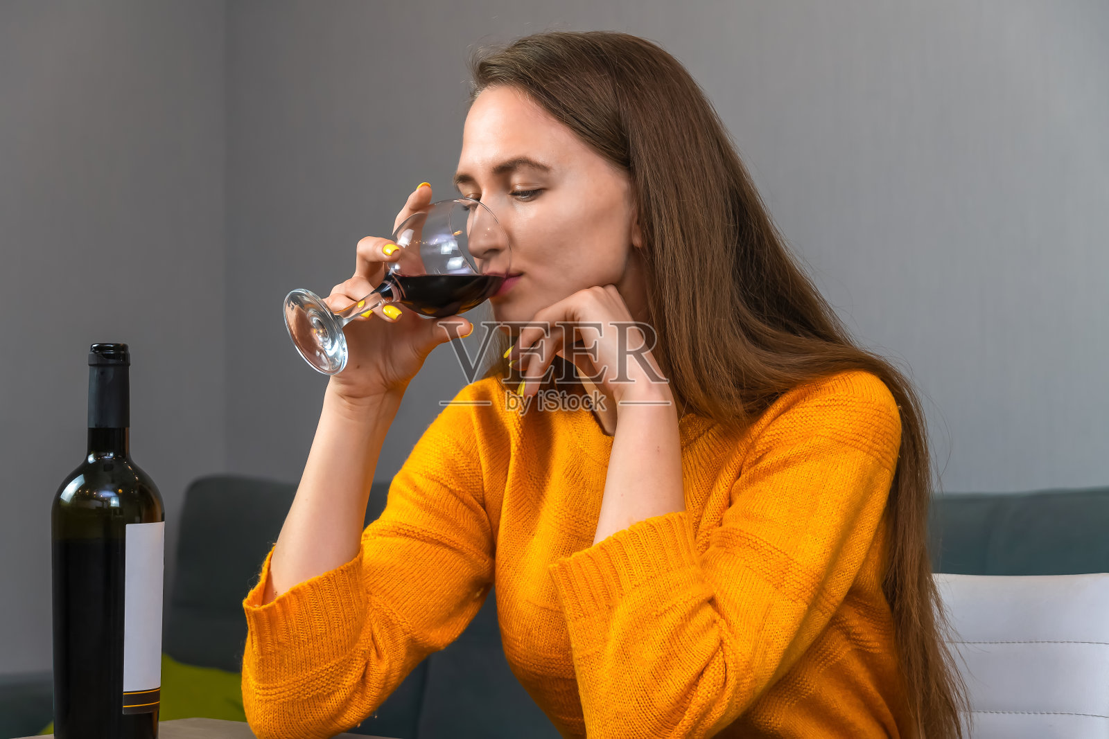 漂亮的年轻黑发女人穿着橙色的运动衫坐在客厅的一张大桌子旁，喝着一杯红酒照片摄影图片