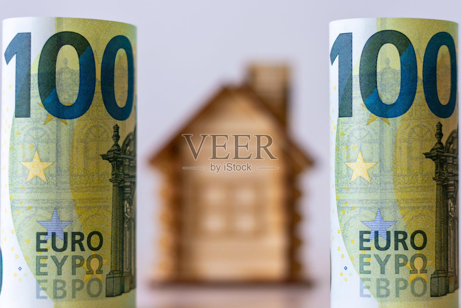 边上是两张100欧元的钞票，卷成一卷。中间有一座隐约可见的房子。房地产市场预算的概念。投资建设。照片摄影图片